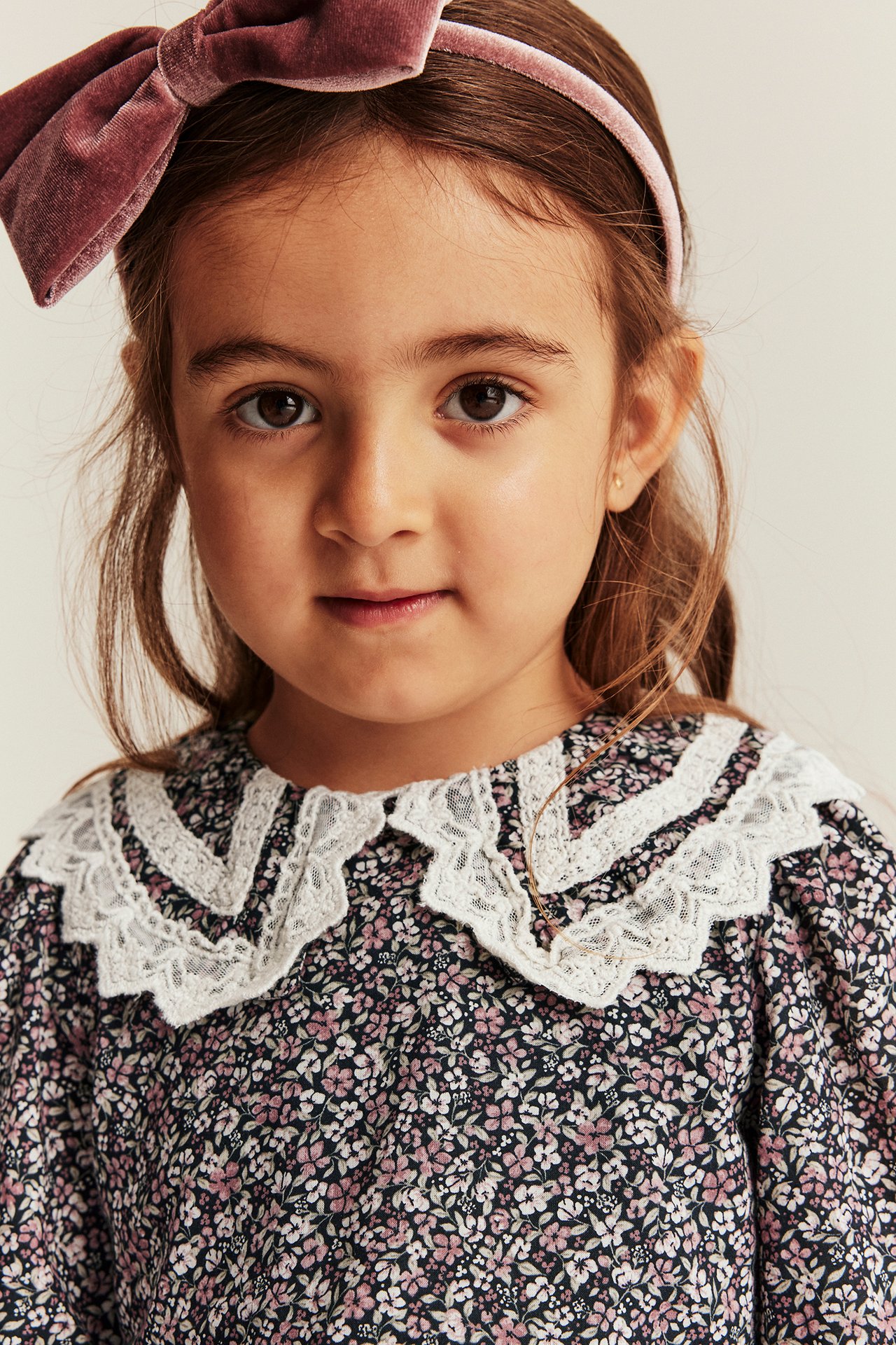 Nahaufnahme: Kind in dunkel geblümtem Baumwollkleid mit weißem Spitzenkragen und rosa Samtstirnband.