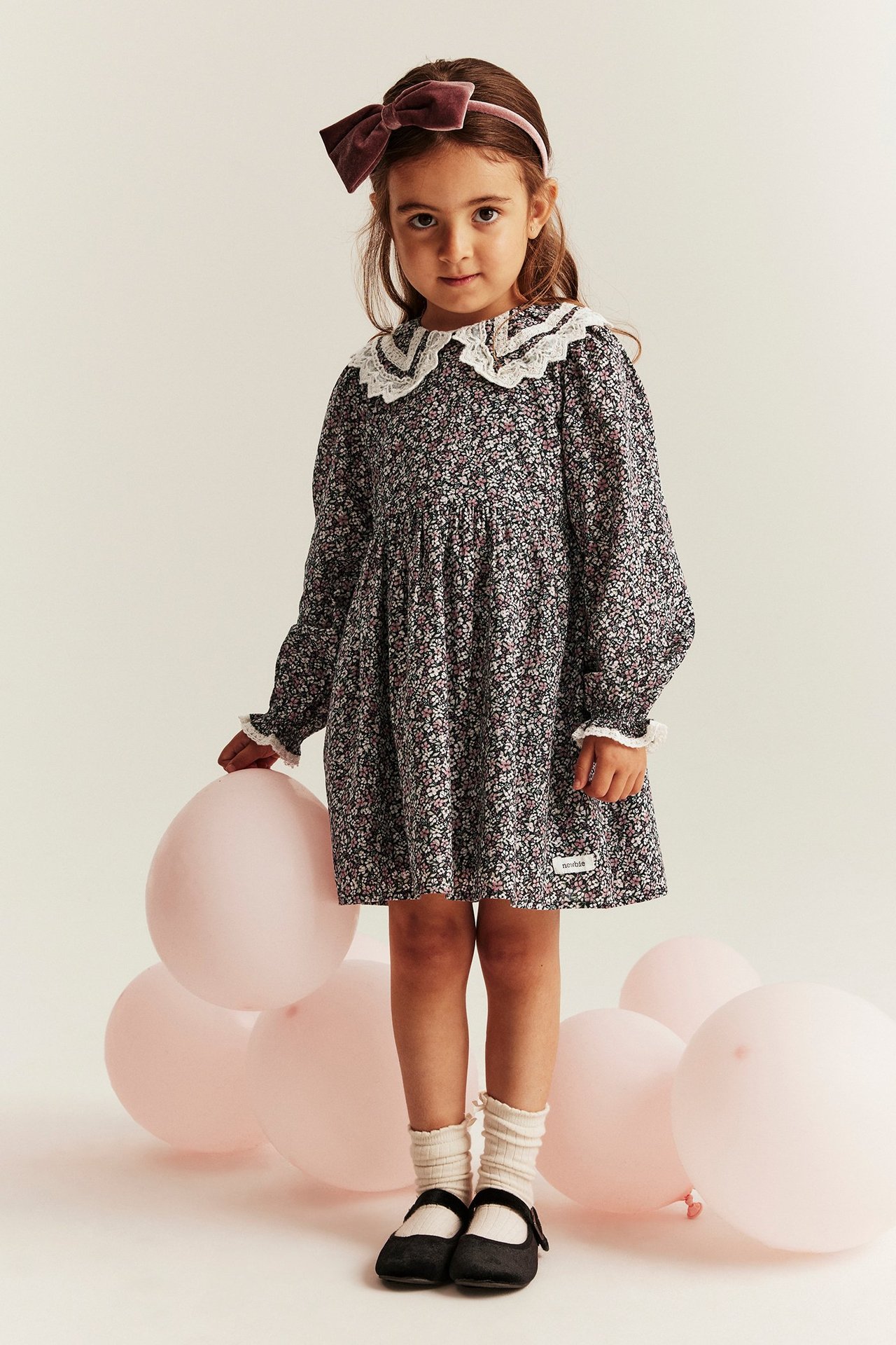 Front view: Kid in a black floral cotton dress with white lace collar, a pink bow headband.