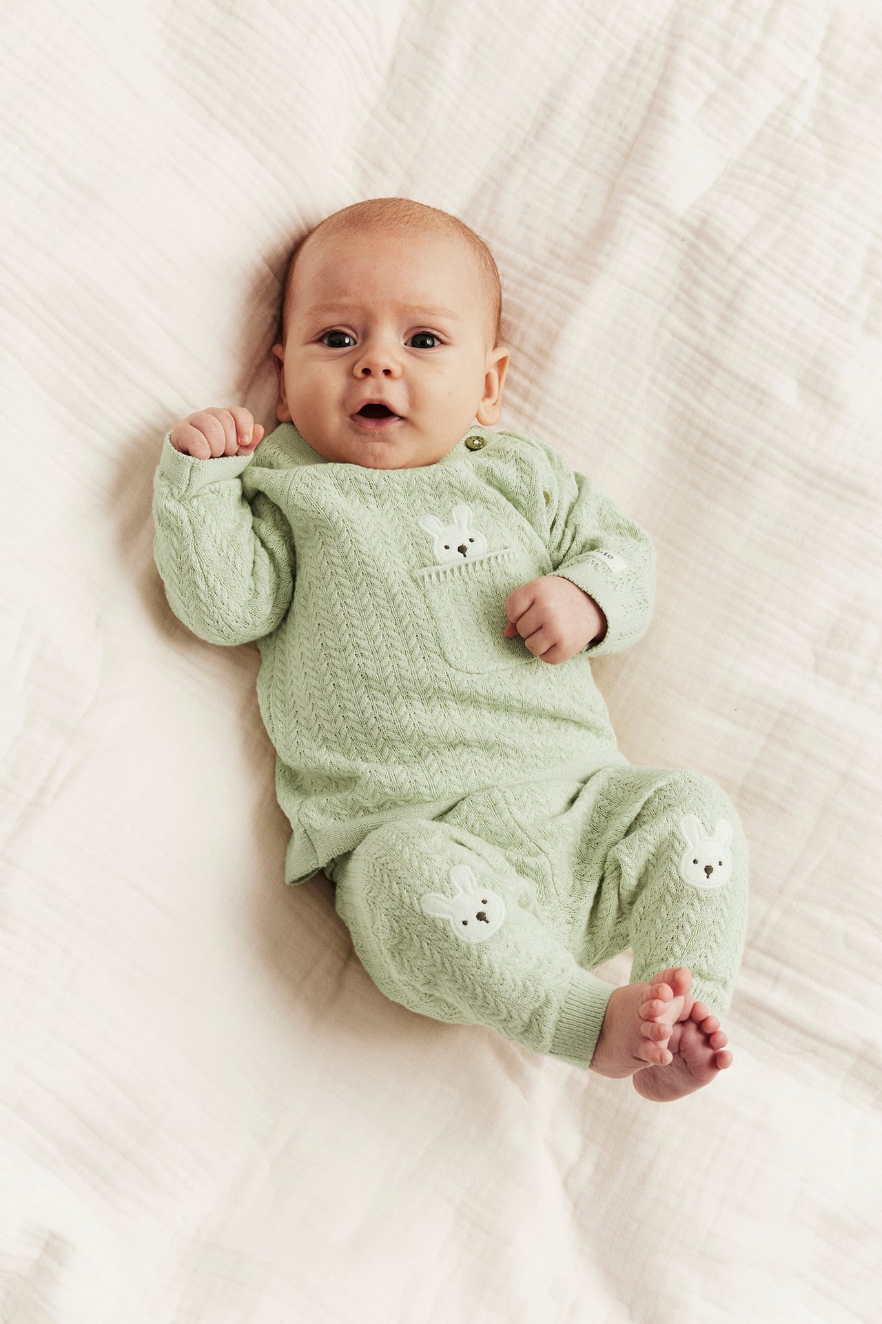 Front view: Baby in light green structured knit top with bunny pocket, shoulder buttons, and matching bunny-knee trousers.