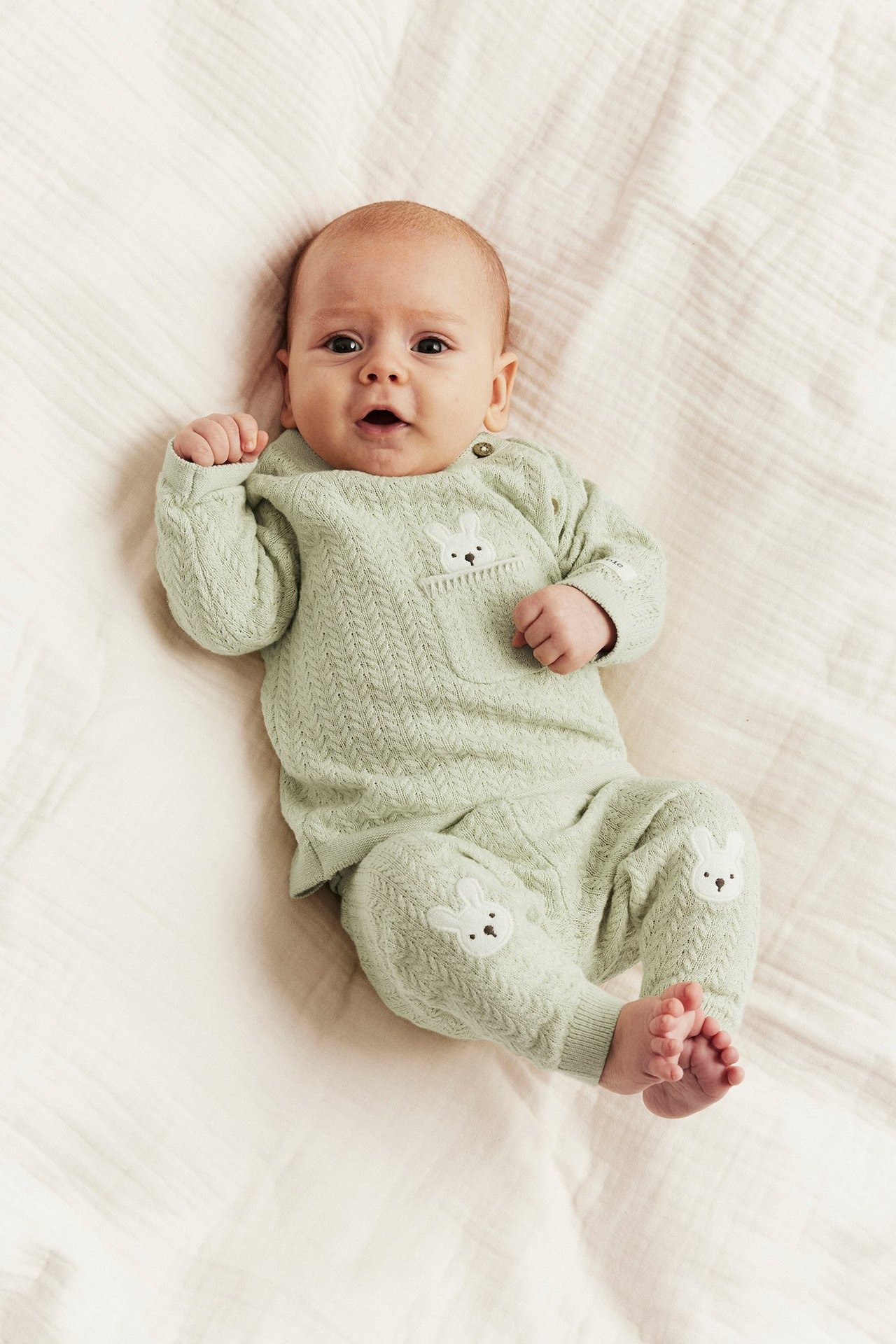 Front view: baby in light green structured knit cotton cardigan with bunny pocket and matching bunny-knee trousers.
