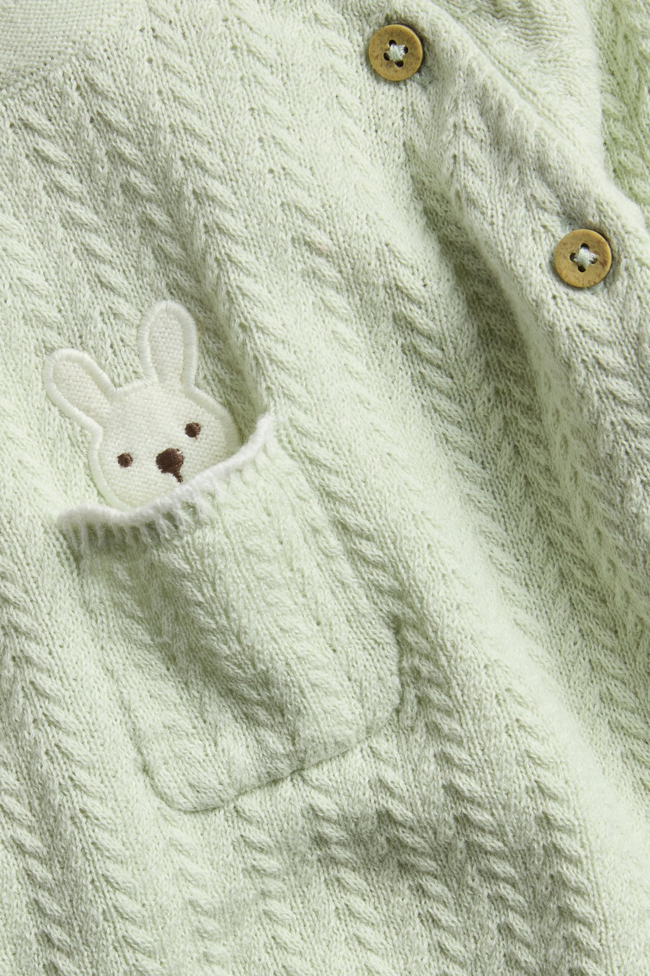 Close-up: Light green structured knit baby cardigan with bunny embroidery pocket and wooden buttons.
