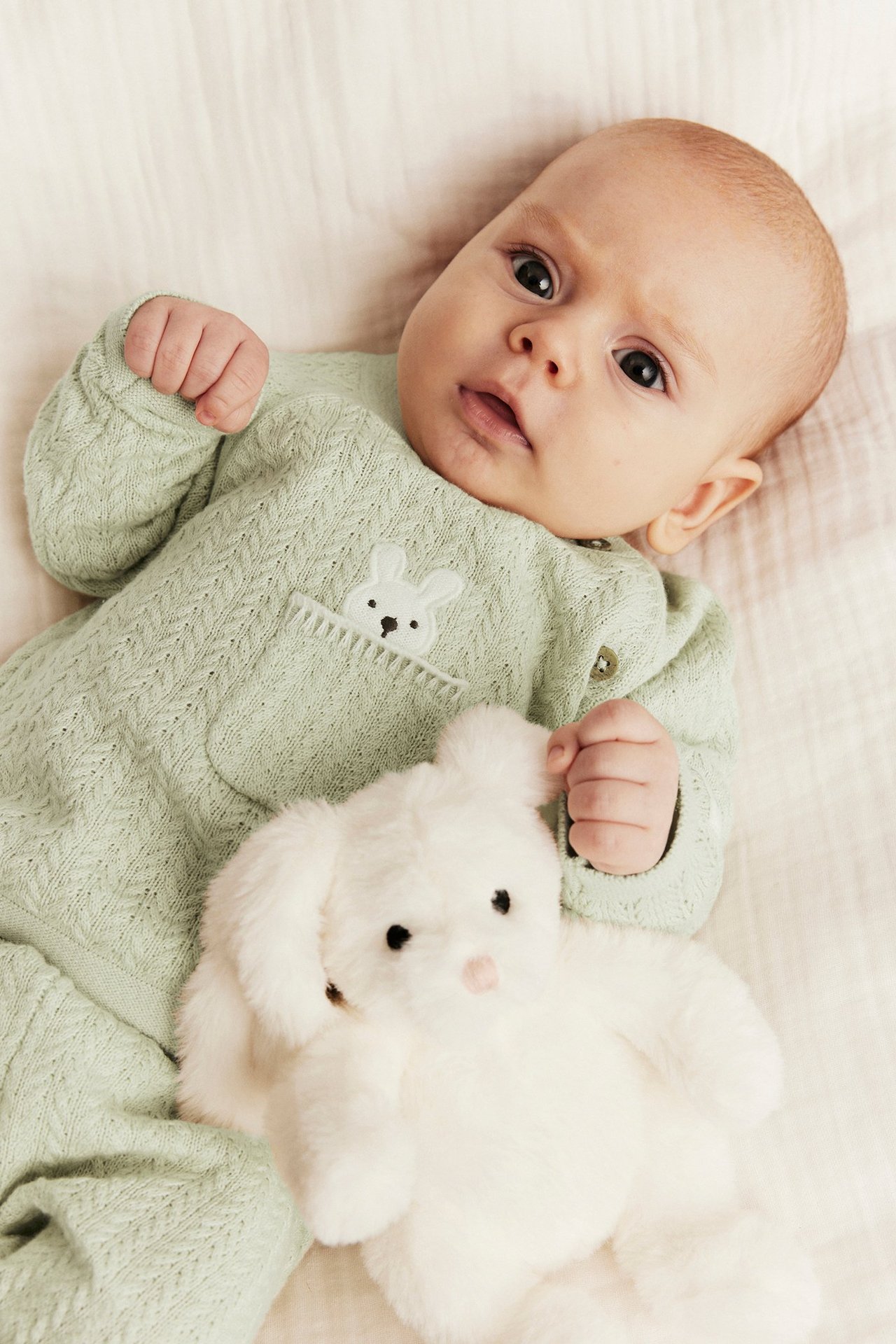 Baby wears a light green patterned knit cotton cardigan with a bunny pocket, holding a white plush bunny.