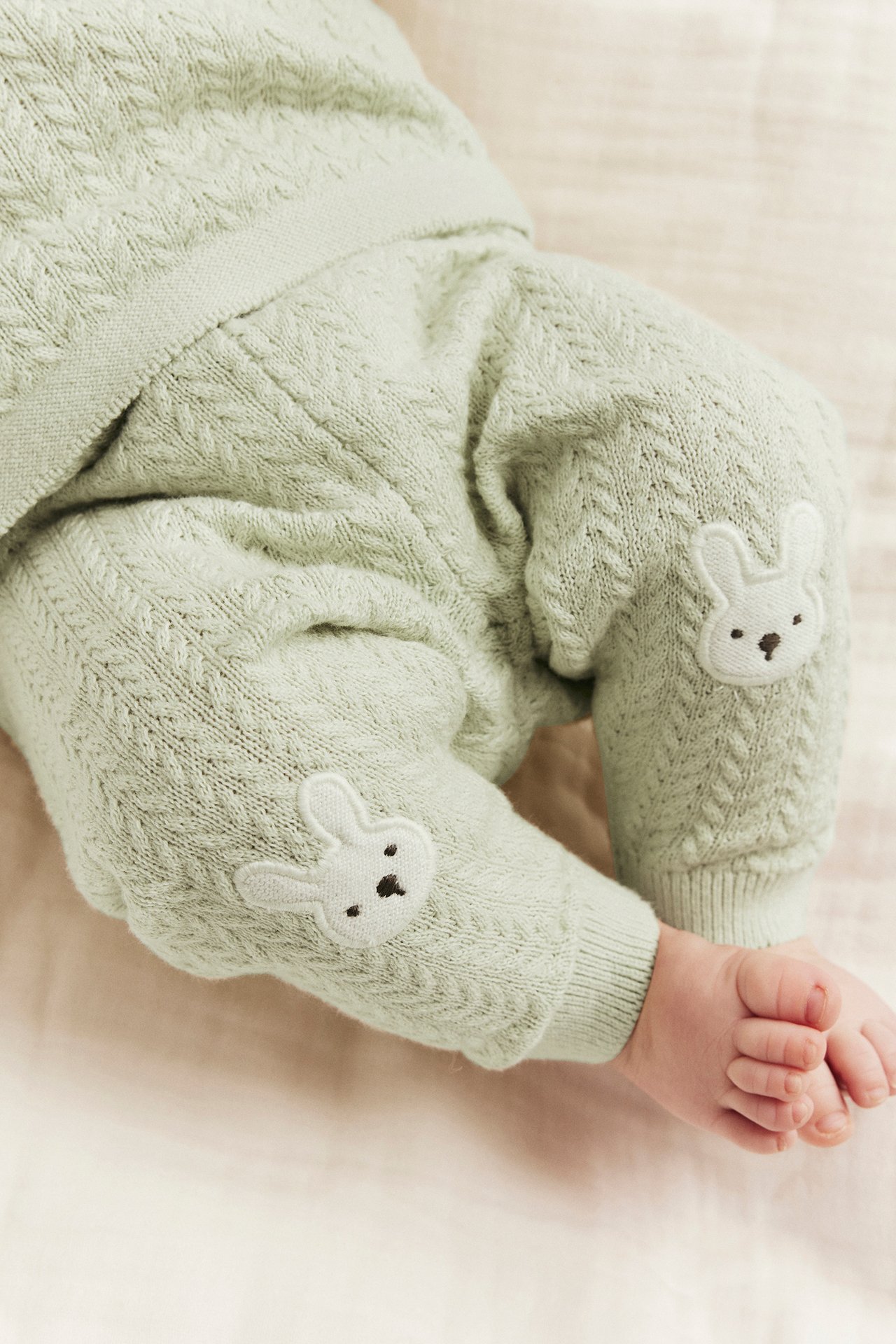 Nahaufnahme eines Babys in hellgrünen, strukturgestrickten Leggings mit weißen Hasenapplikationen an den Knien.