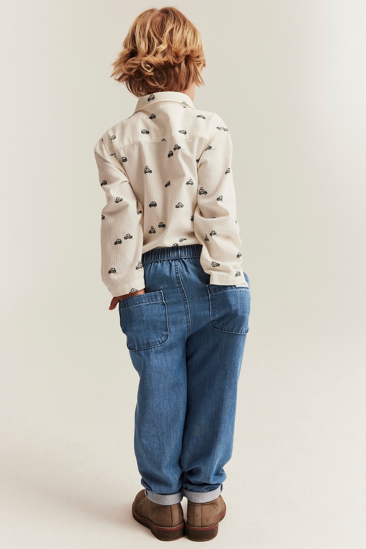Back view of a kid wearing blue denim trousers with an elasticated waist and a light car-patterned shirt.