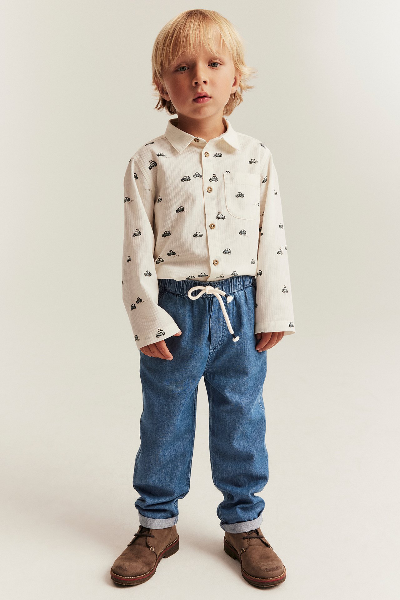 Front view of a kid wearing blue denim trousers with a drawstring and a cream car-patterned shirt.