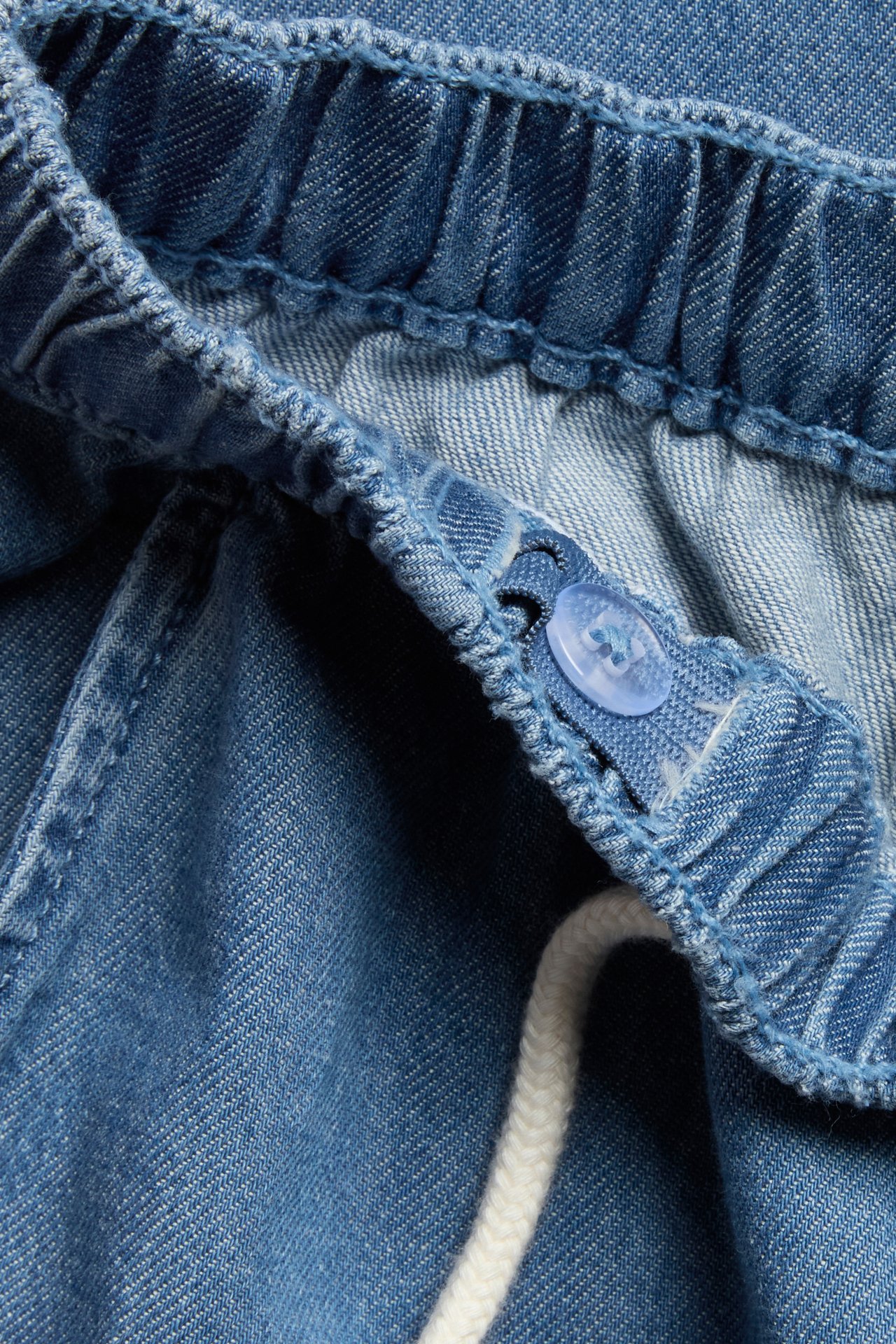 Close-up of blue denim trousers' elastic waistband with a white drawstring and a light blue button.
