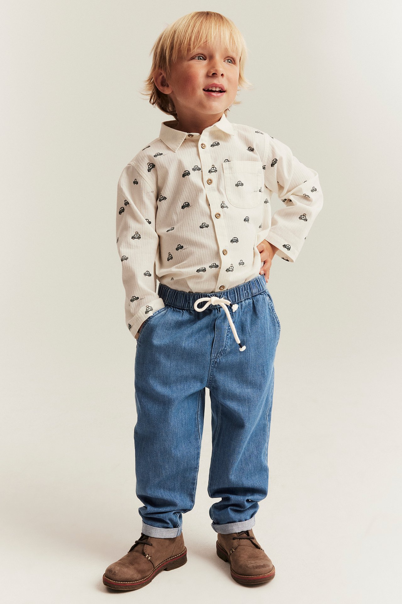 Front view of a kid wearing blue denim drawstring trousers, a cream car-patterned shirt, and brown shoes.
