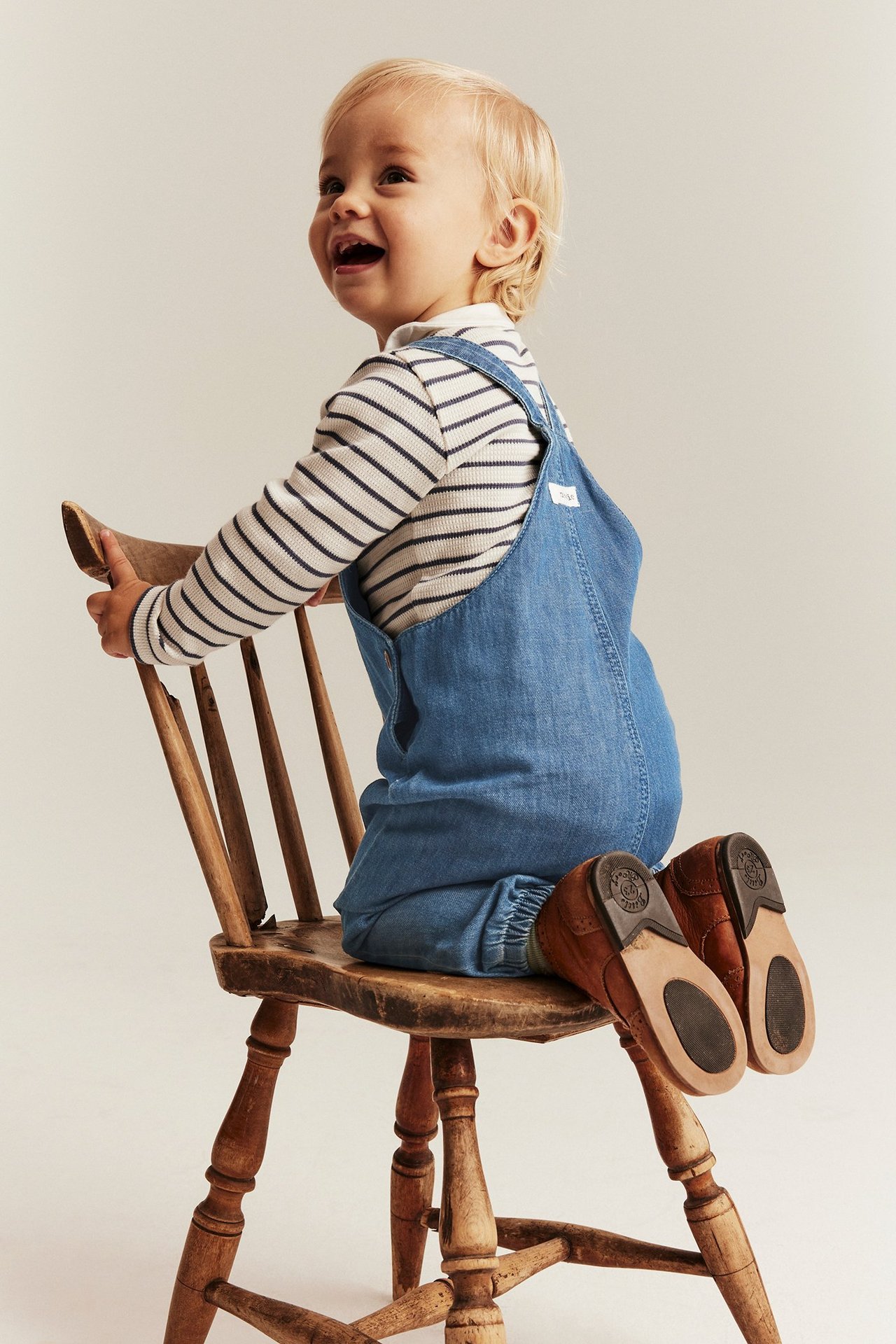 Seitenansicht eines Babys in hellblauer Jeans-Latzhose mit Gummibündchen, einem gestreiften Oberteil und braunen Schuhen.