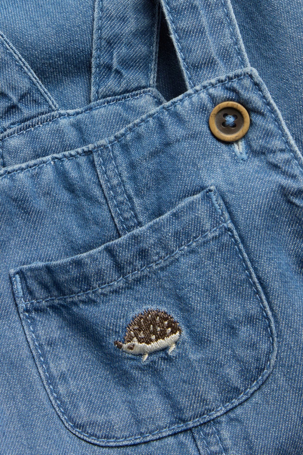Close-up of baby's blue denim dungarees with hedgehog embroidery on the chest pocket and a wooden button.