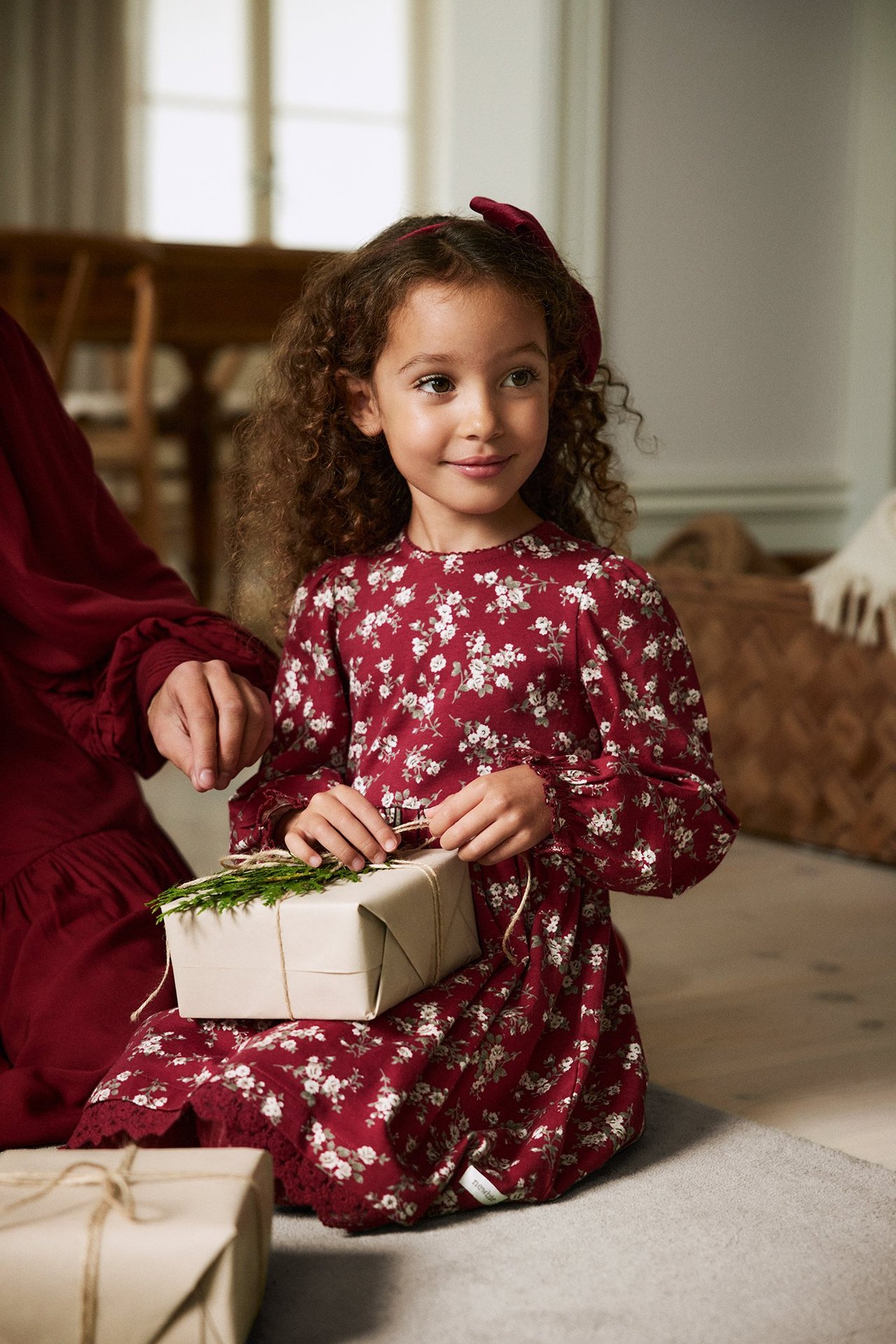 Front-side view: Girl in red floral jersey dress with lace hem, red bow, holding gift.