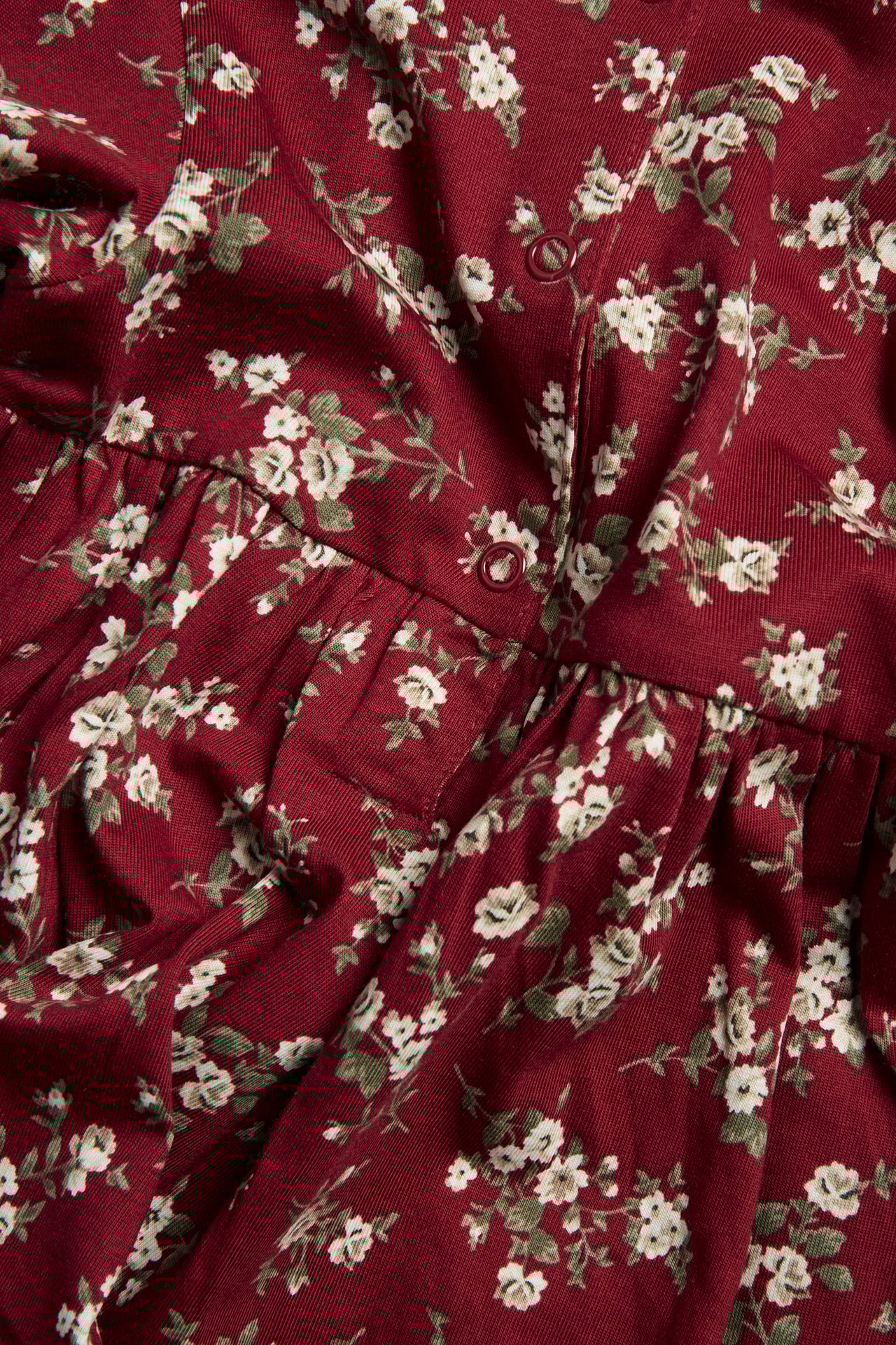 Nahaufnahme eines roten Jersey-Kinderkleides mit weißem Blumenmuster, roten Druckknöpfen und geraffter Taille.