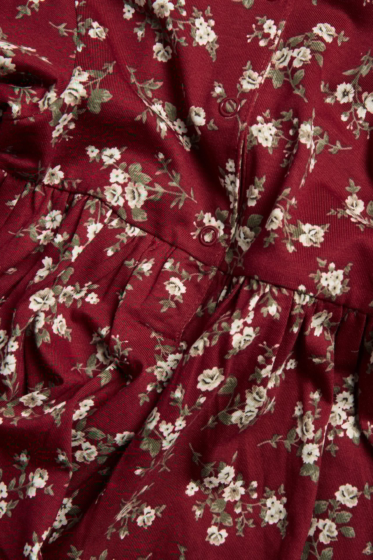 Close-up of a red jersey kid's dress with a white floral print, featuring red snap buttons and a gathered waist.