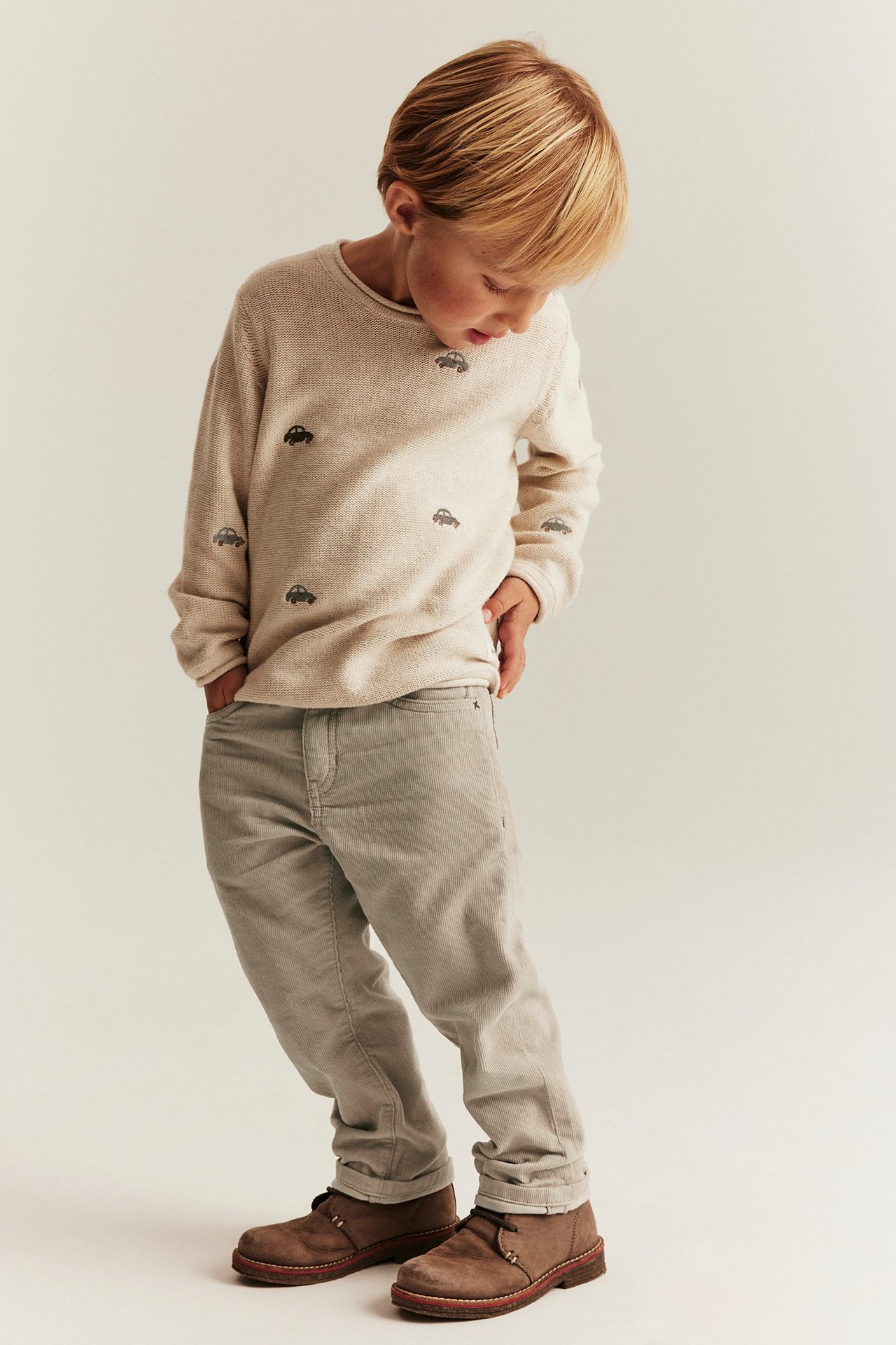 Vorderansicht Kind in beiger Cordhose, einem gestrickten Auto-Pullover und braunen Stiefeln.