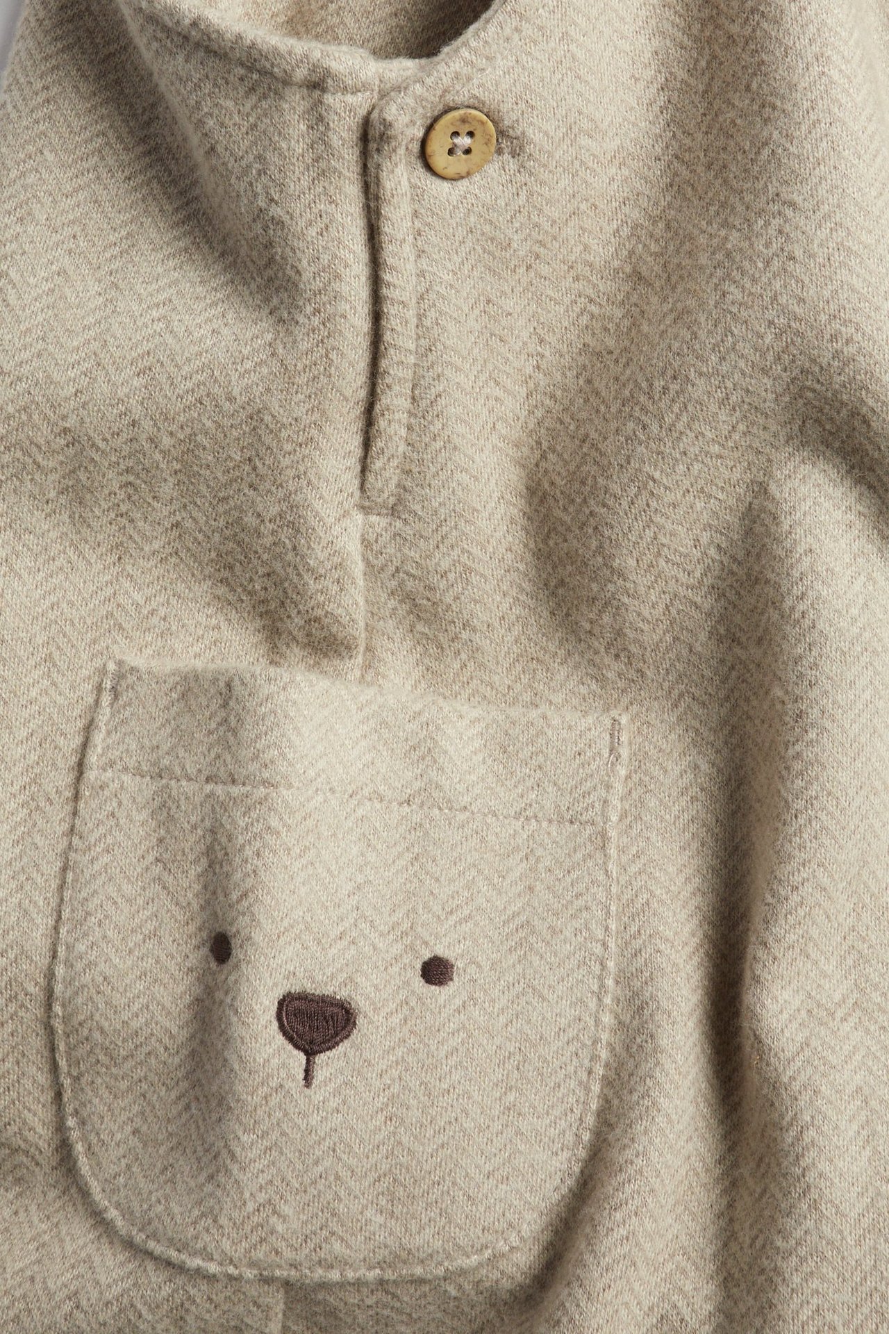 Close-up of beige melange herringbone overalls with a wooden button and a pocket with bear face embroidery.