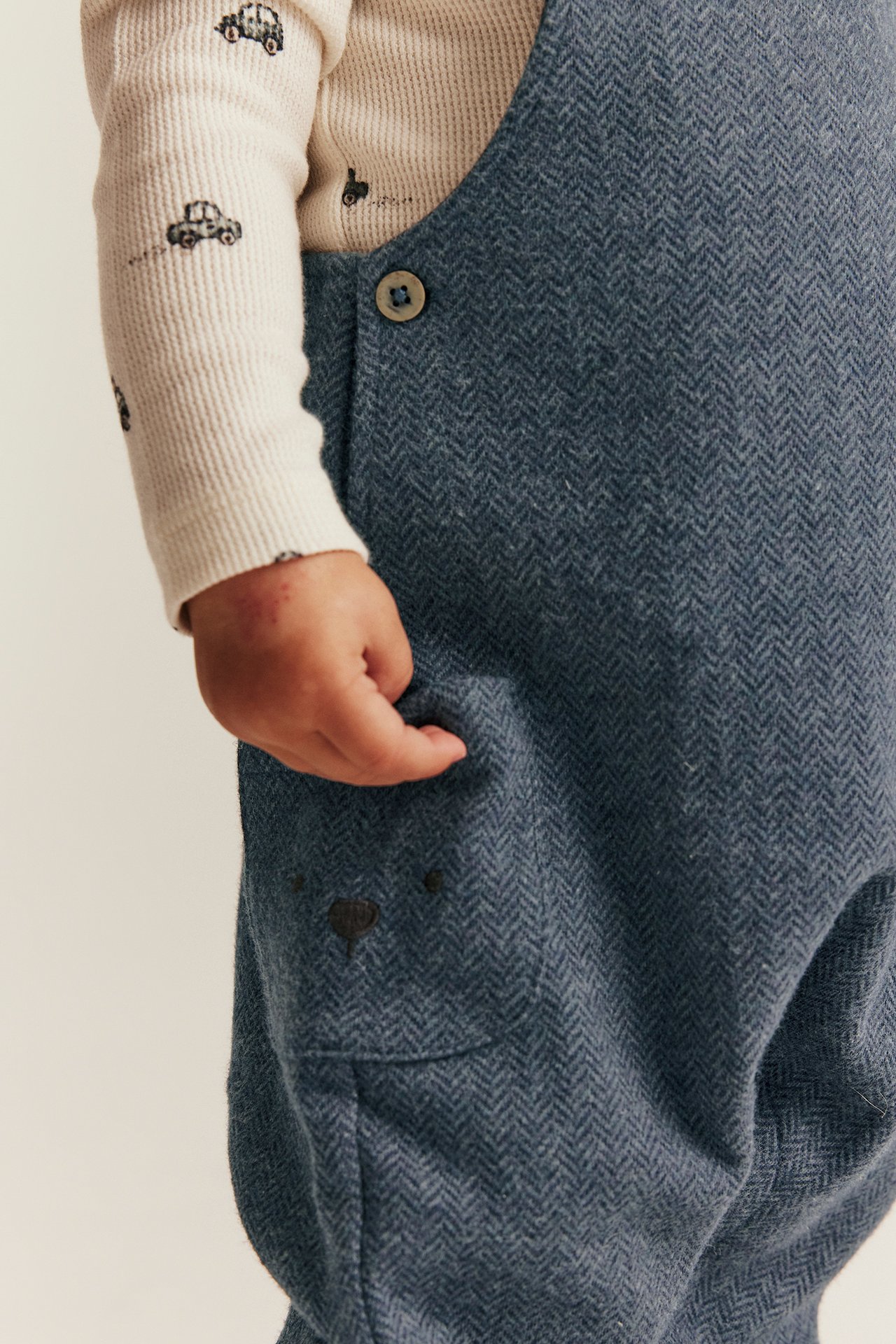 Baby wears blue herringbone dungarees with embroidered bear, over cream car-print long-sleeve top.