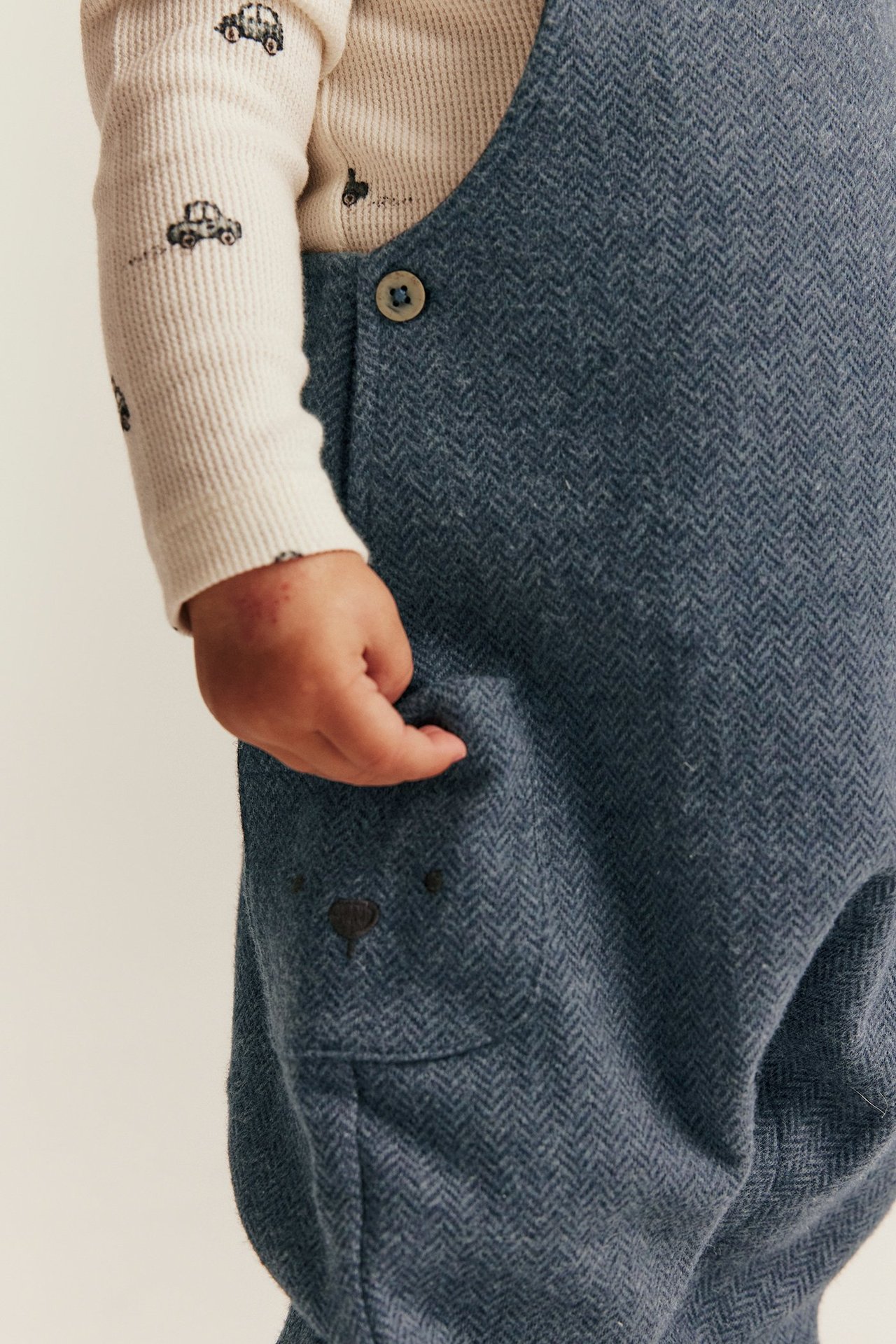 Baby trägt blaue Fischgrät-Latzhose mit gesticktem Bären über einem cremefarbenen Langarmshirt mit Automotiven.