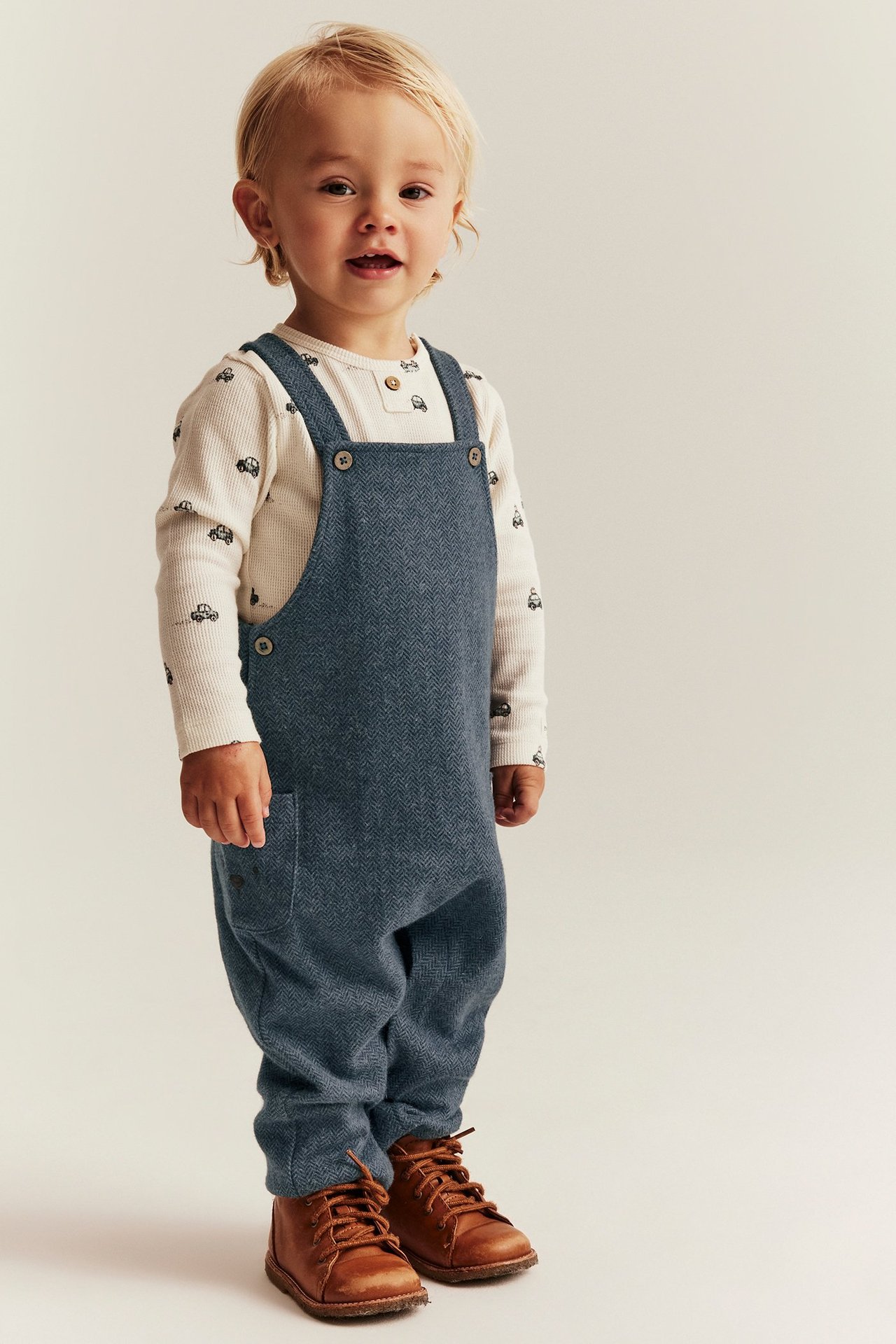 Vorderansicht Baby: blaue Jacquard-Latzhose mit Fischgrätmuster, weißes Auto-Shirt, braune Schnürstiefel.