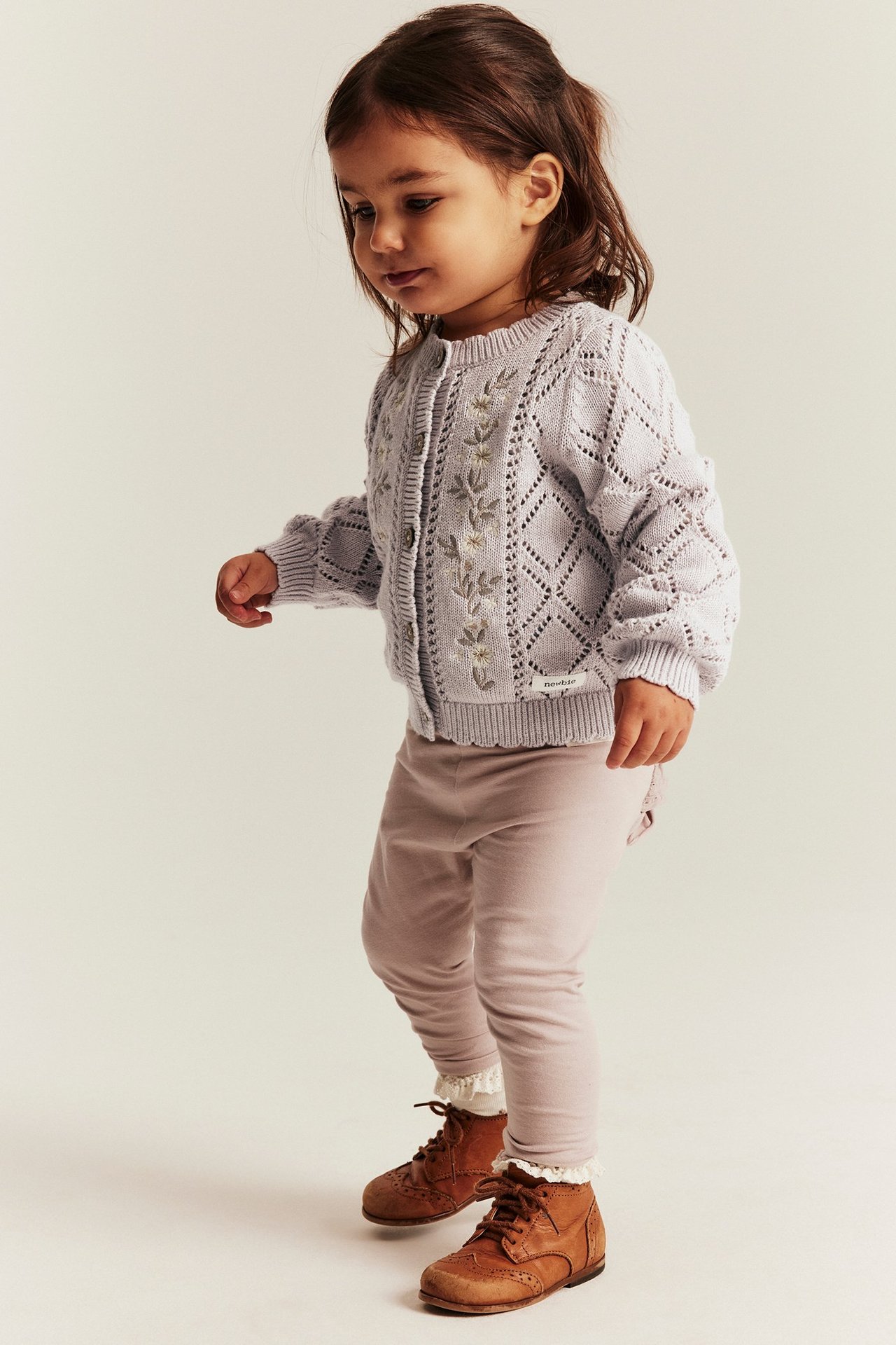 Vorderansicht eines Babymädchens in hellfliederfarbener Strickjacke mit Blumenstickerei, rosa Leggings und braunen Stiefeln.