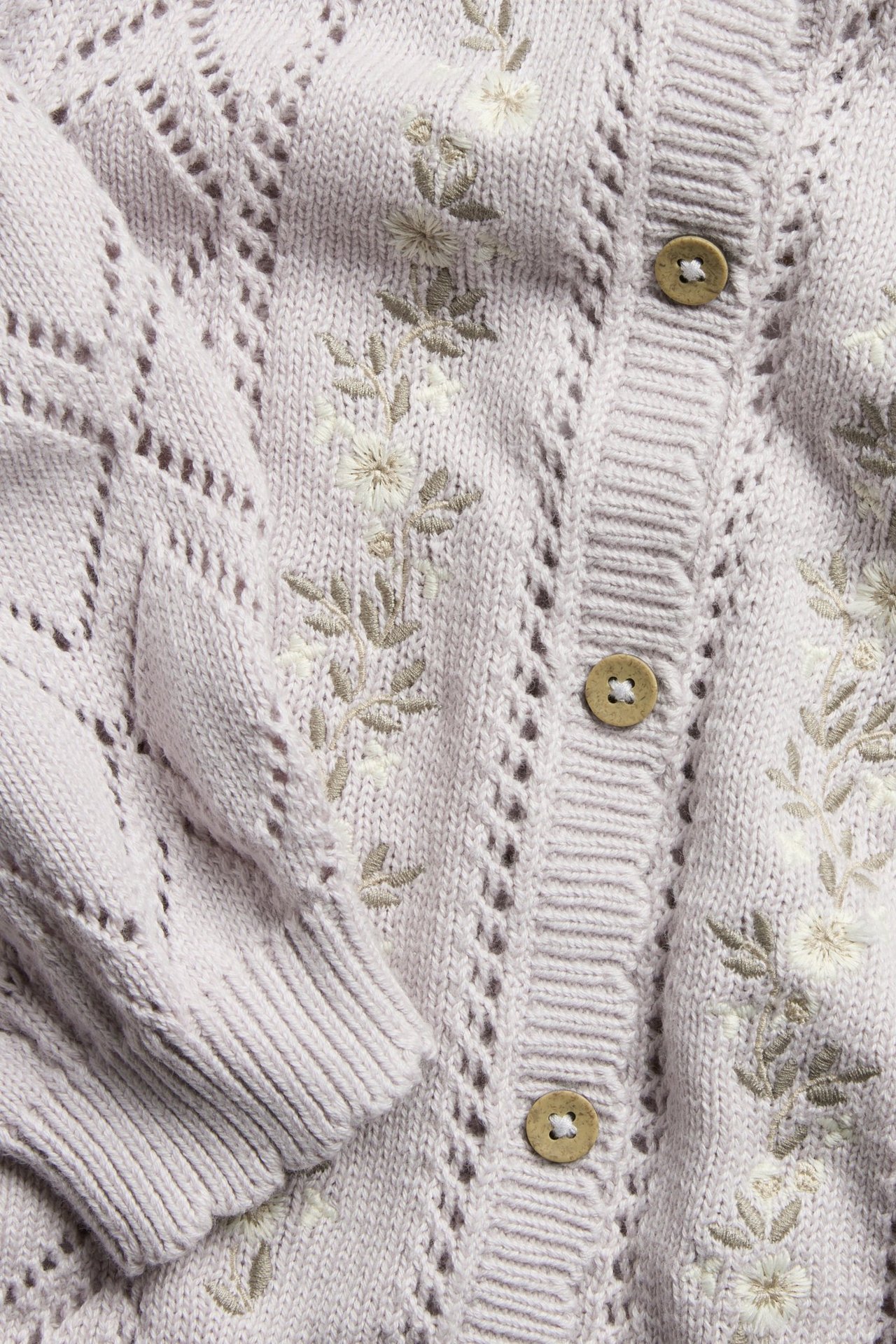 Nahaufnahme einer helllila gemusterten Baumwollstrickjacke mit gestickten Blumen und Holzknöpfen.