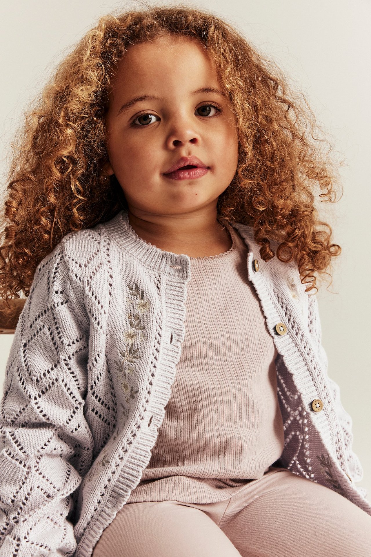 Vorderansicht: Kind in helllila gemusterter Baumwollstrickjacke mit Blumenstickerei über rosa Rippentop und Leggings.