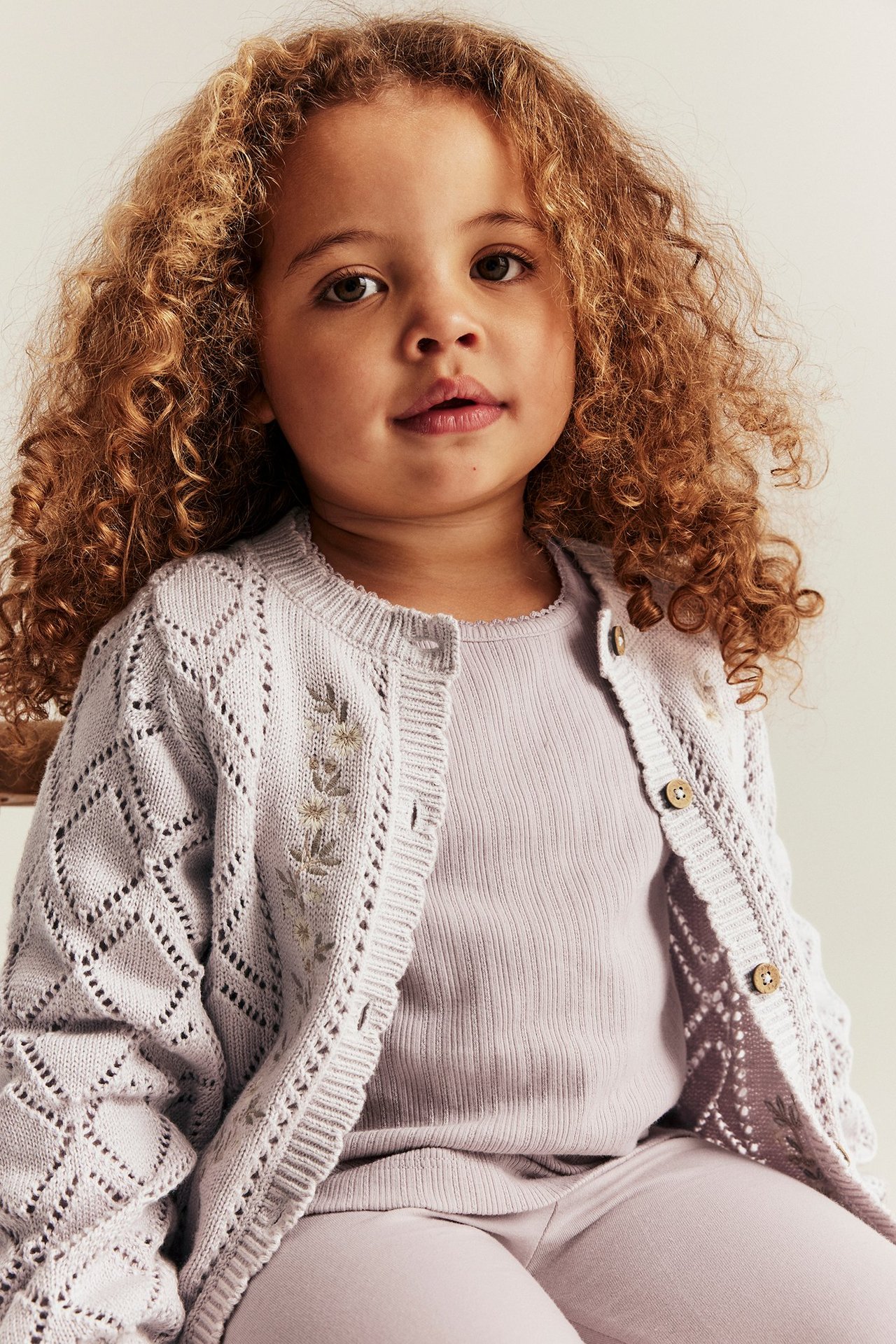 Vorderansicht: Kind in hellfliederfarbener gemusterter Strickjacke aus Baumwolle mit Stickblumen, über passendem Ripp-Top und Leggings.