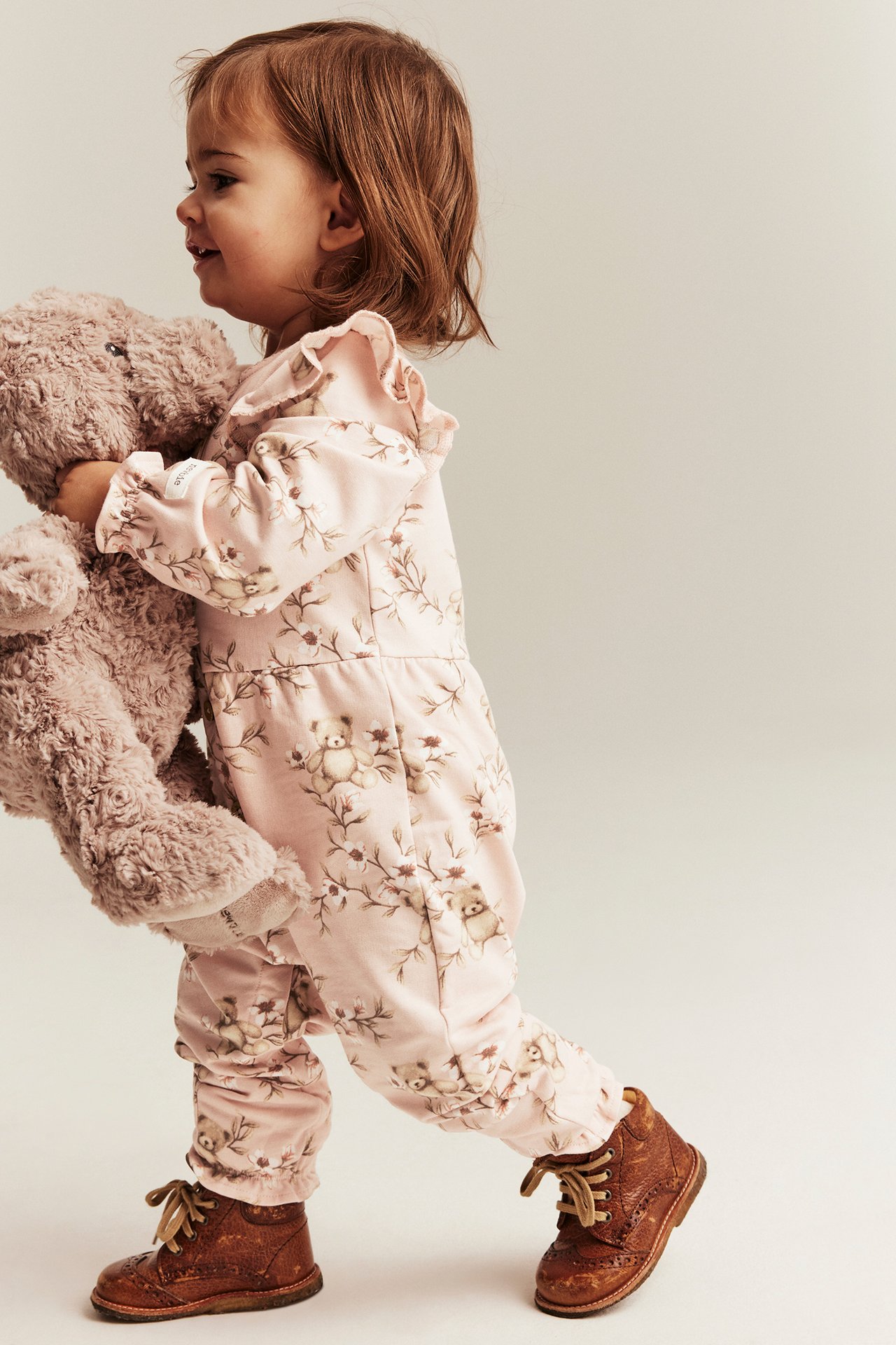 Side view: baby wears light pink cotton floral-teddy jumpsuit with ruffles and brown boots, holding a teddy bear.