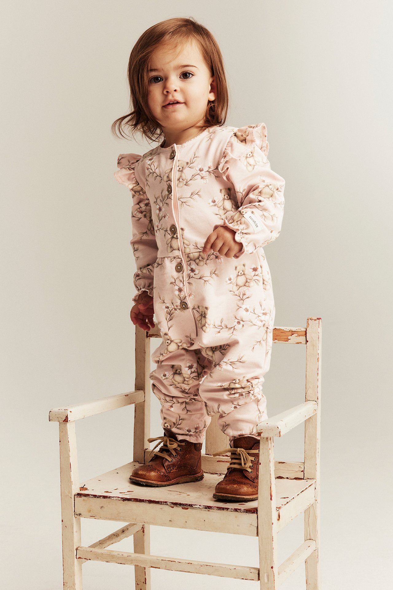 Front view of a baby wearing a light pink floral cotton jumpsuit with shoulder ruffles and brown lace-up boots.