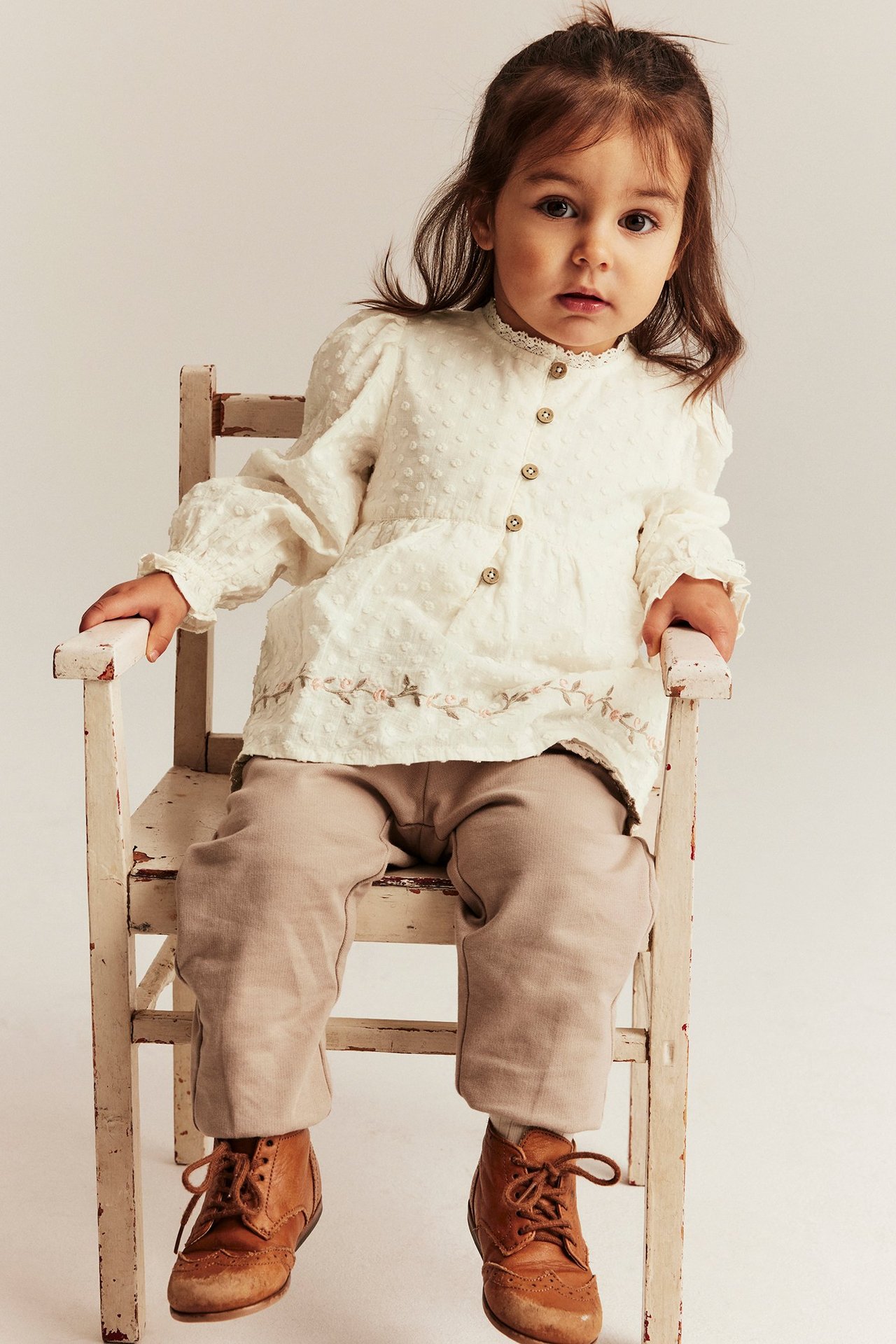 Off-white textured blouse bodysuit with floral embroidery, light brown pants, and brown boots on a kid.