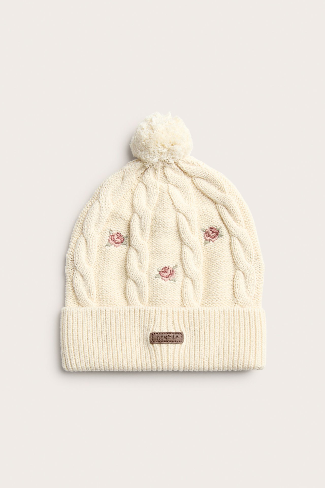 Front view: Light beige knitted baby/kid beanie with cable knit, pink embroidered roses, ribbed brim, pom-pom.