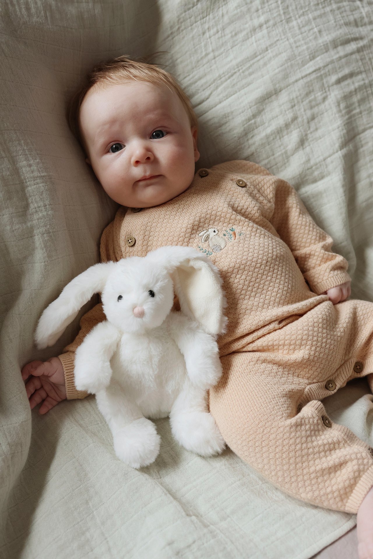Vorderansicht: Baby trägt hellorangefarbenen strukturgestrickten Overall mit Kaninchenstickerei, neben einem weißen Plüschhasen.