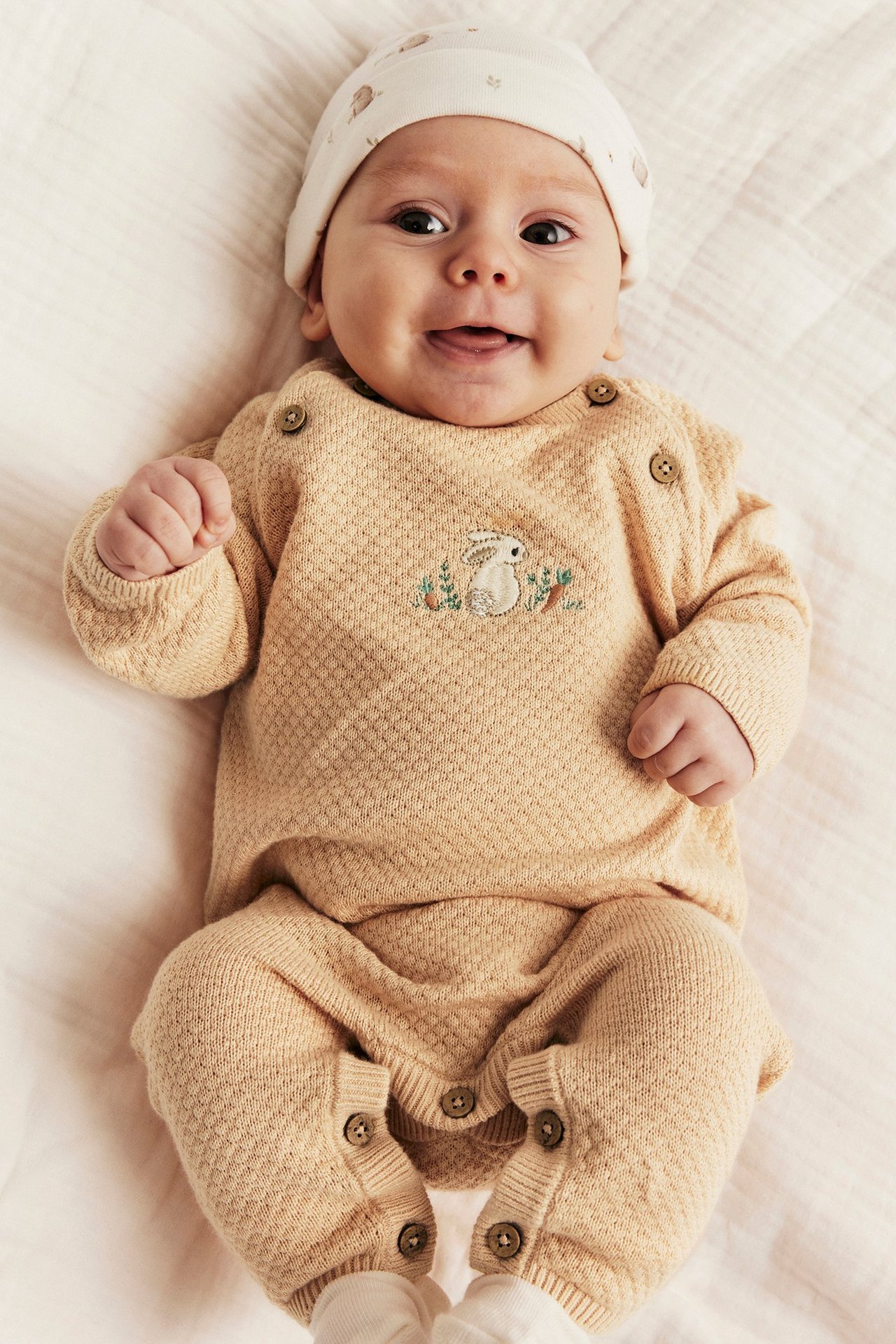 Front view: baby in a light orange structured knit coverall with rabbit embroidery and a patterned hat.