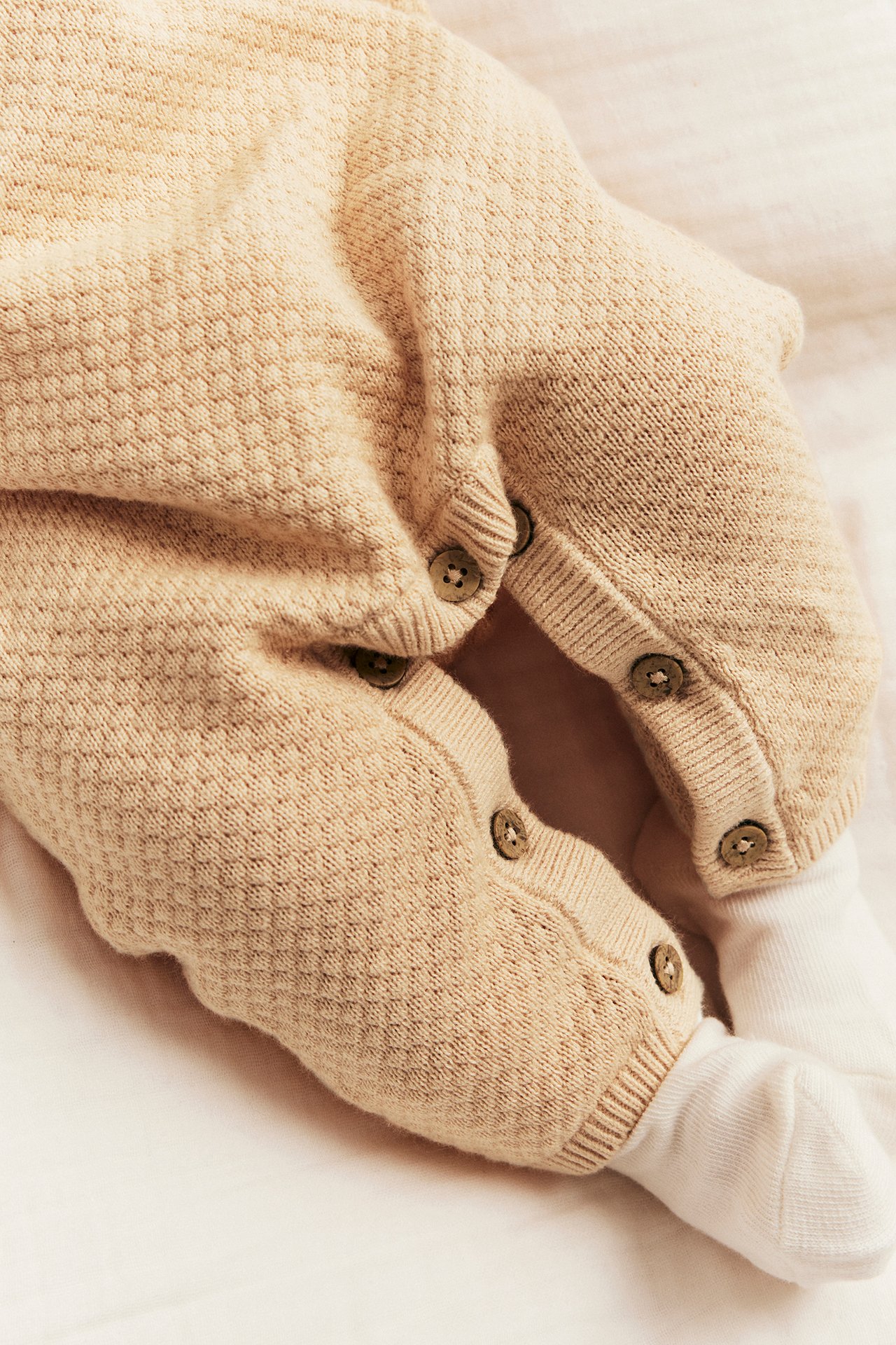 Close-up of a baby's legs wearing a light orange structured knit cotton jumpsuit with buttons, styled over white socks.