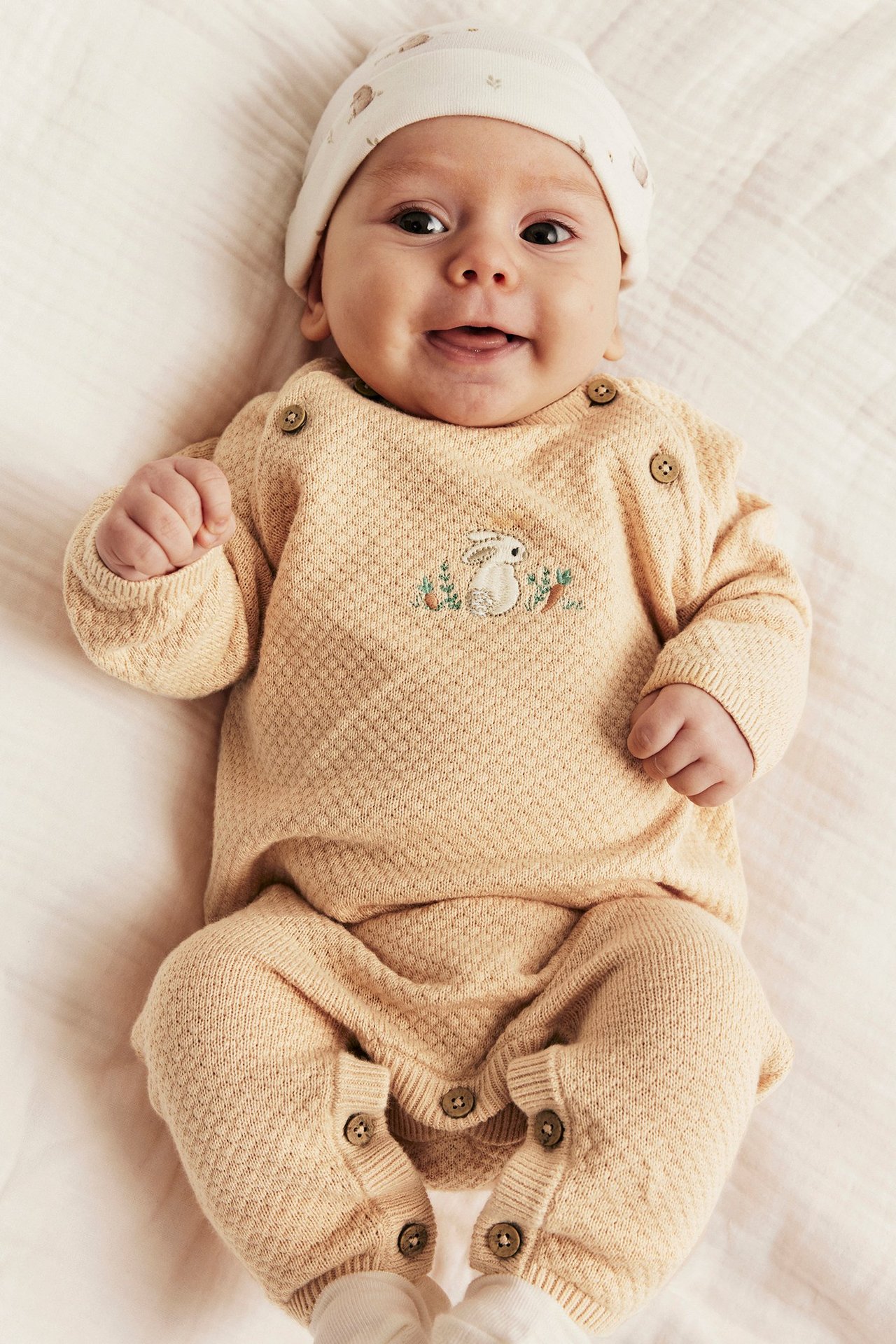 Front view of a baby in a light orange structured knit jumpsuit with embroidered rabbit and white patterned beanie.