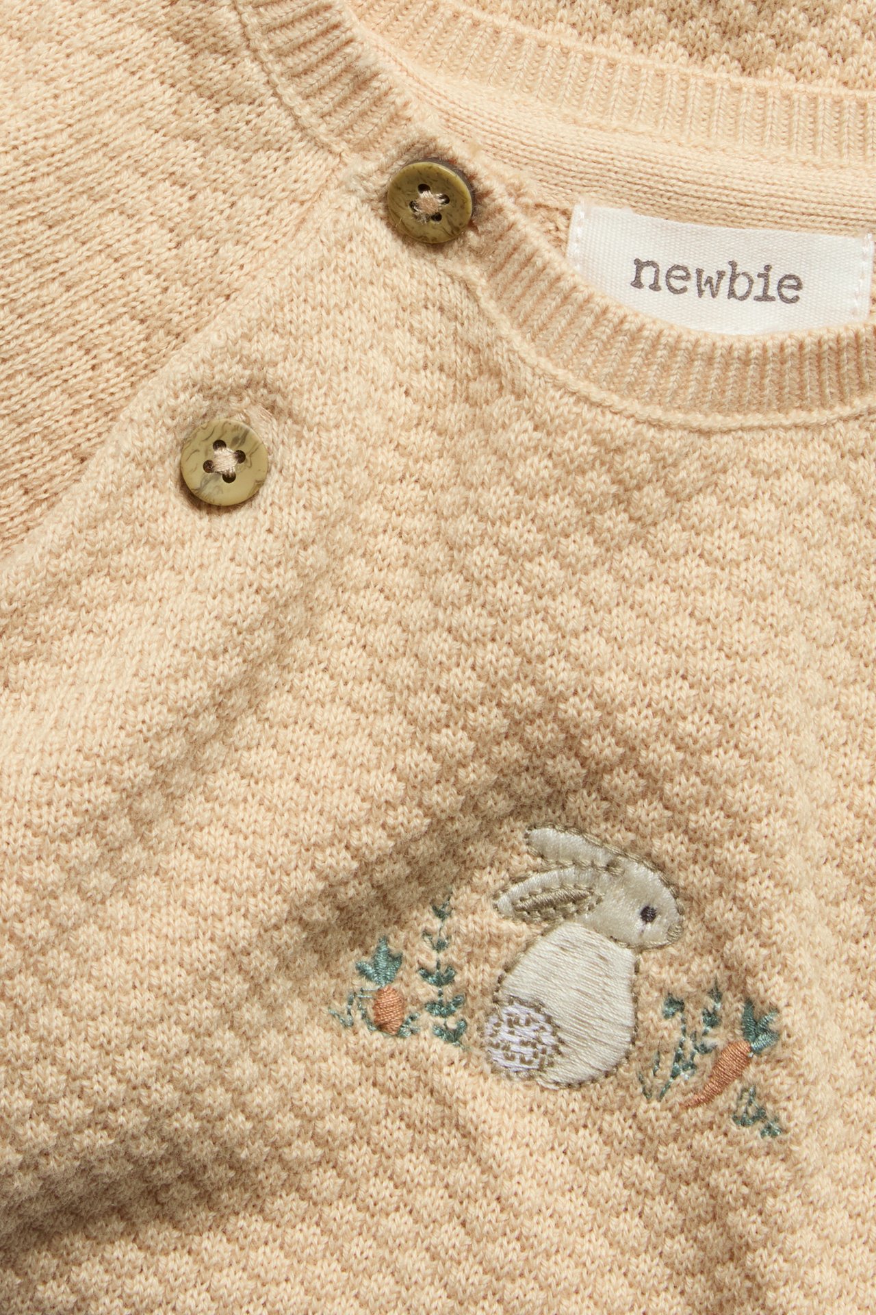 Close-up: light orange structured knit baby jumpsuit with embroidered rabbit, carrots, shoulder buttons.