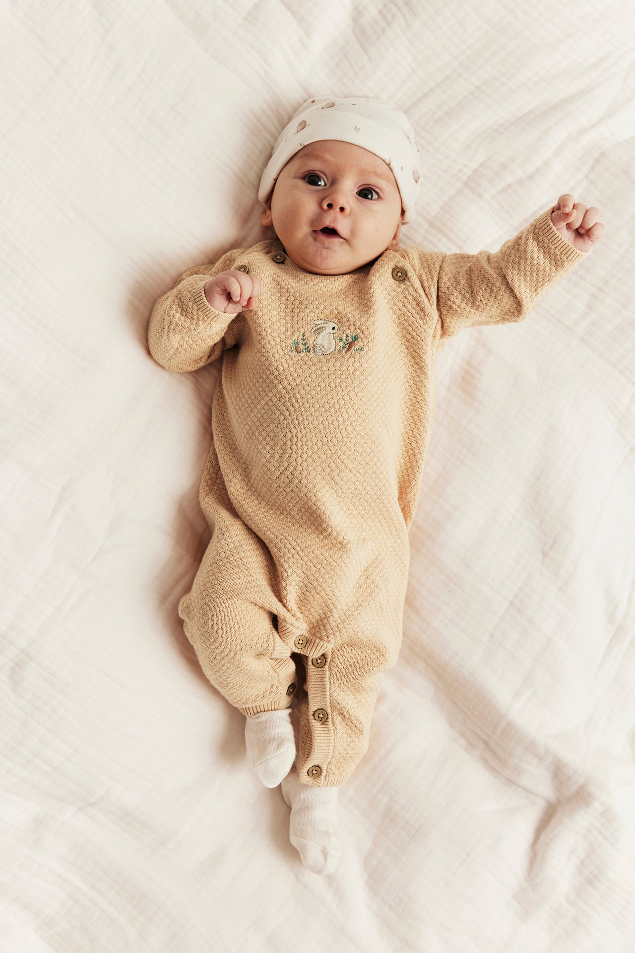 Baby wears light orange knit jumpsuit with rabbit embroidery, white socks, and patterned beanie.