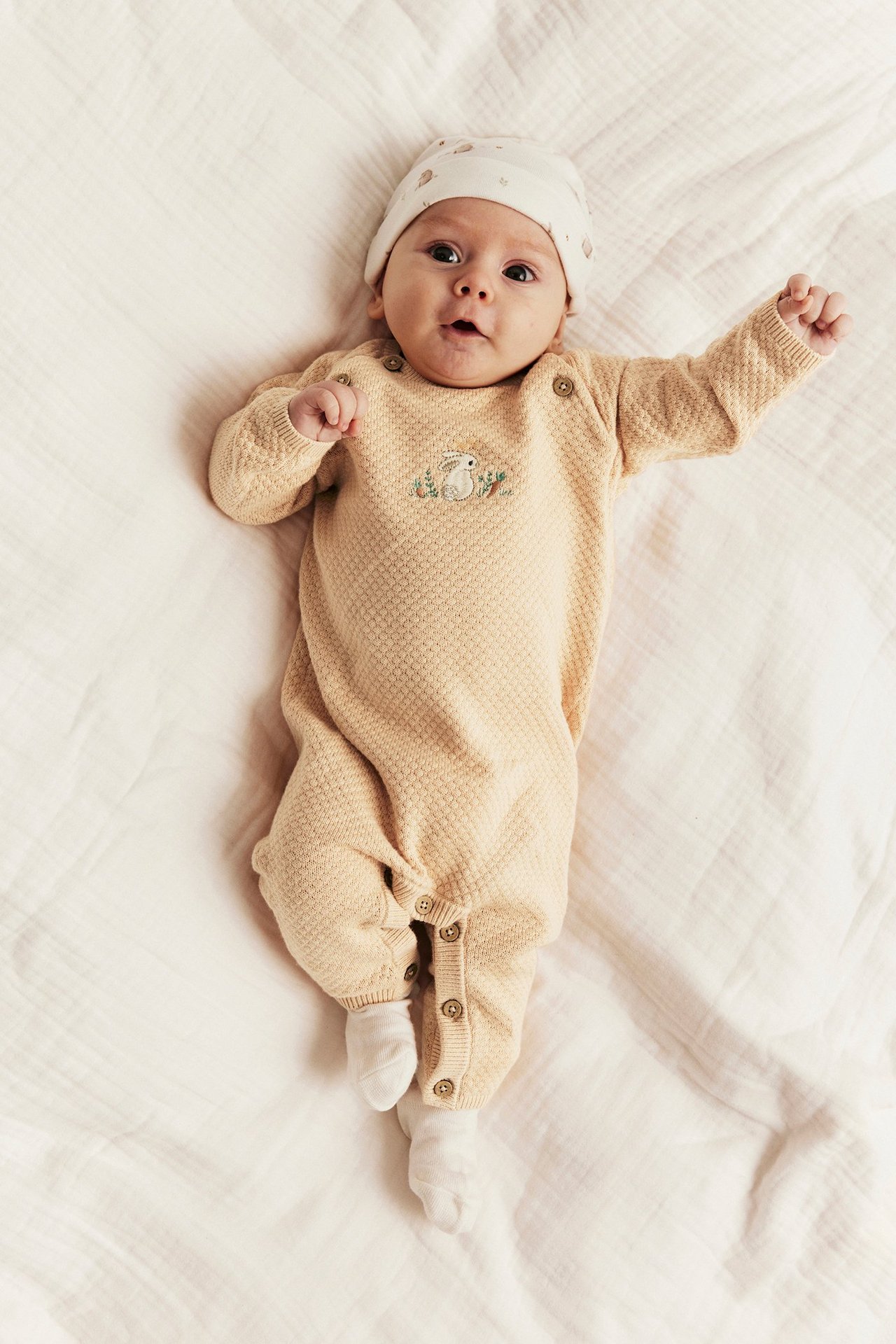 Front view: baby in light beige knit cotton jumpsuit with rabbit embroidery, a patterned hat, and white socks.