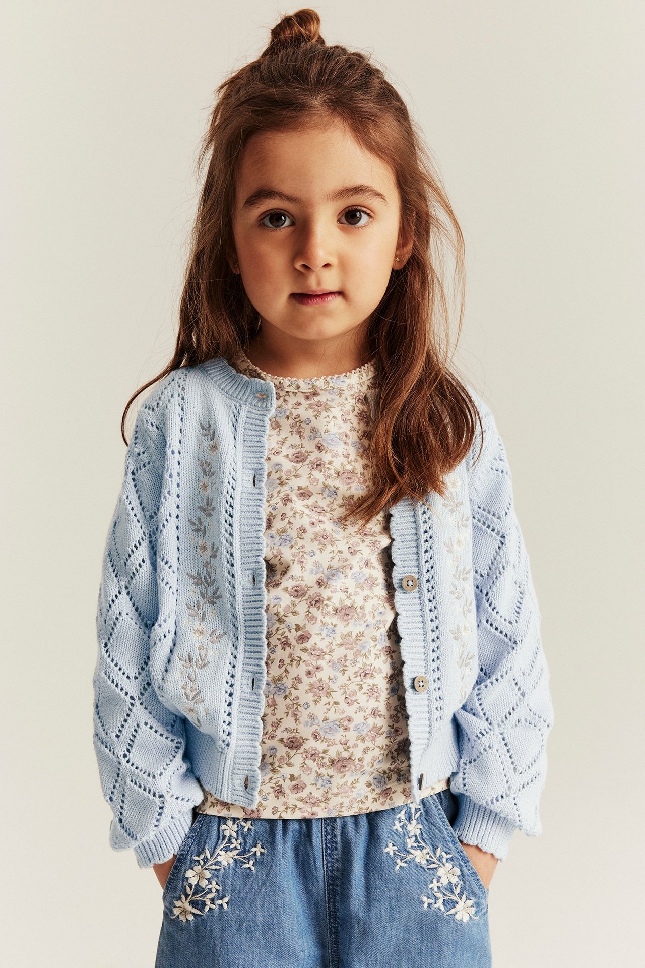 Vorderansicht: Kind in hellblauer bestickter Strickjacke, über cremefarbenem Blumentop und blauen bestickten Shorts.