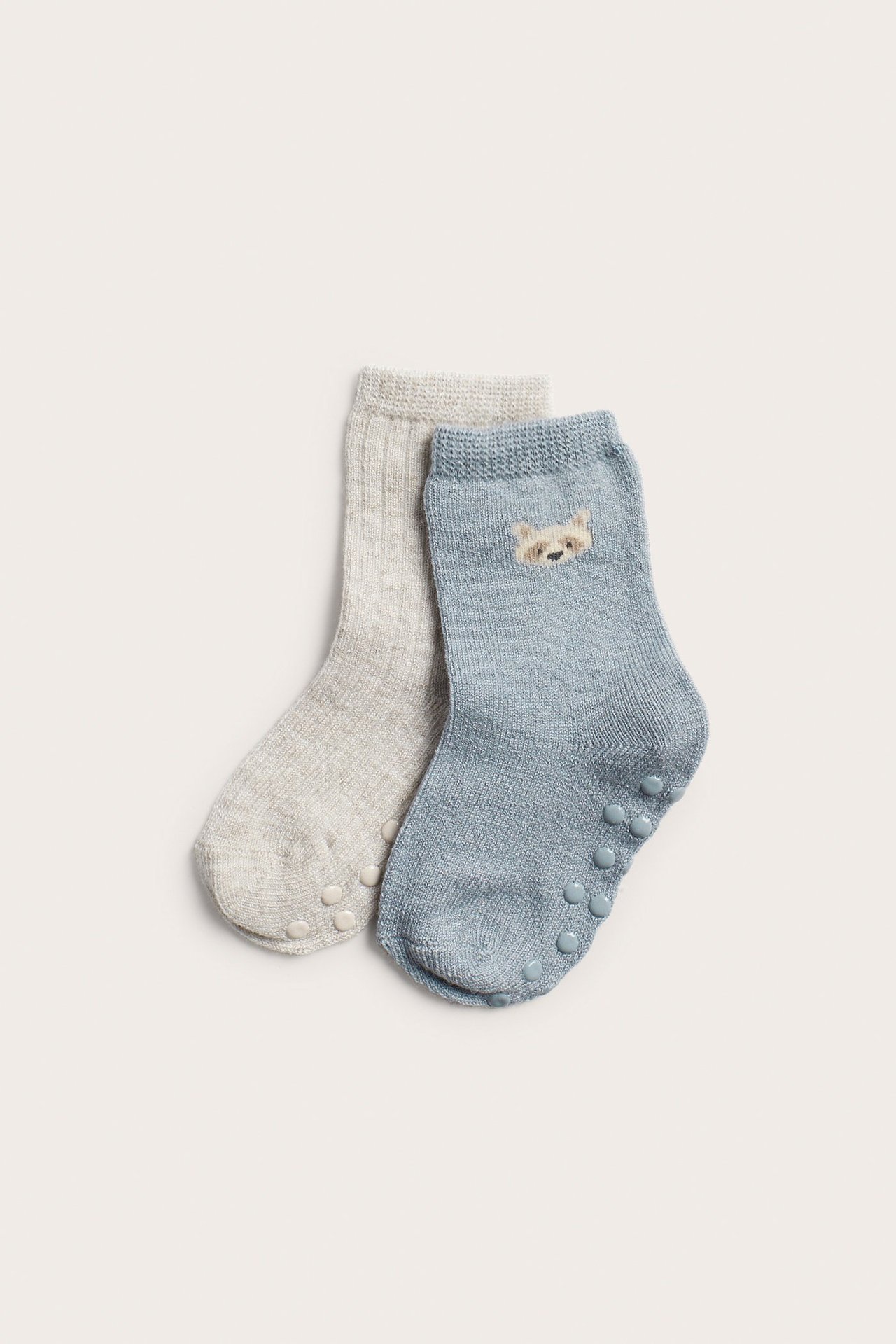 Zwei Baby-Wollmixsocken: hellblau mit Waschbärgesicht, hellgrau gerippt, beide mit Antirutsch-Noppen.