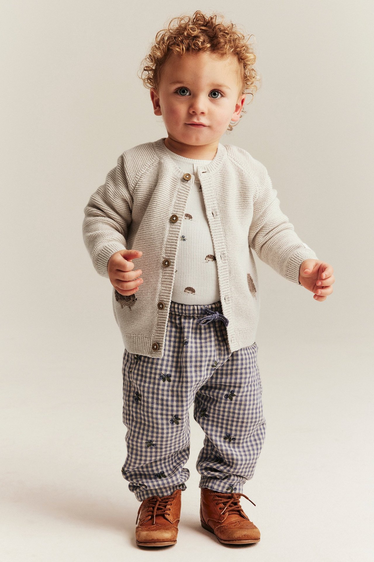Front view: Baby in a beige knitted cardigan with hedgehog embroidery, a light hedgehog print bodysuit, and checked pants.