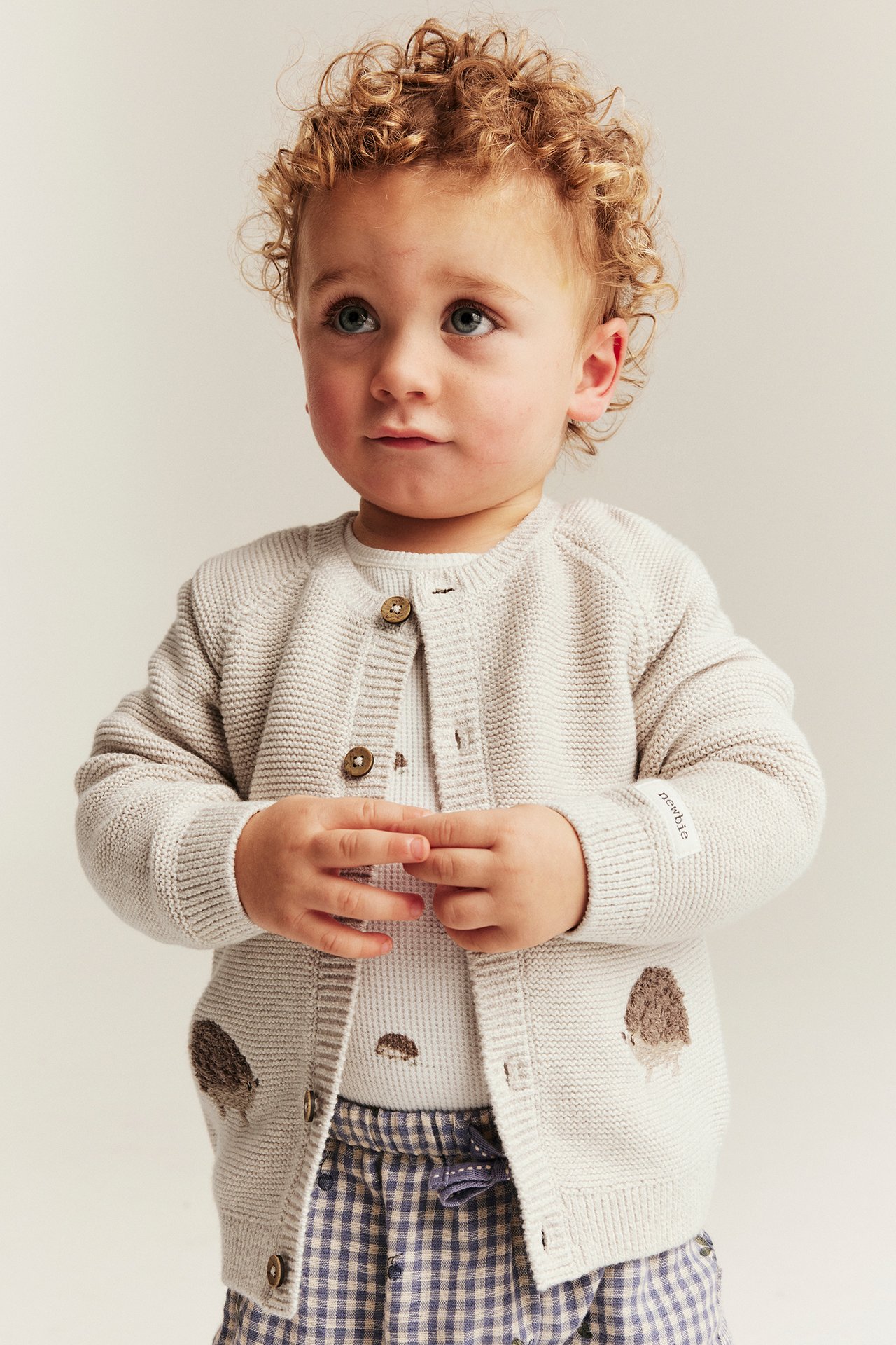 Front view of a baby wearing a beige melange knitted cardigan with hedgehog embroidery over a white top and plaid pants.