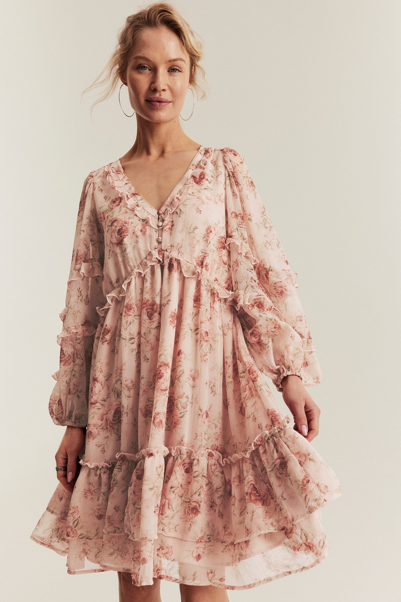 Front view of a woman in a pink floral chiffon dress with ruffle tiers, front buttons, and hoop earrings.