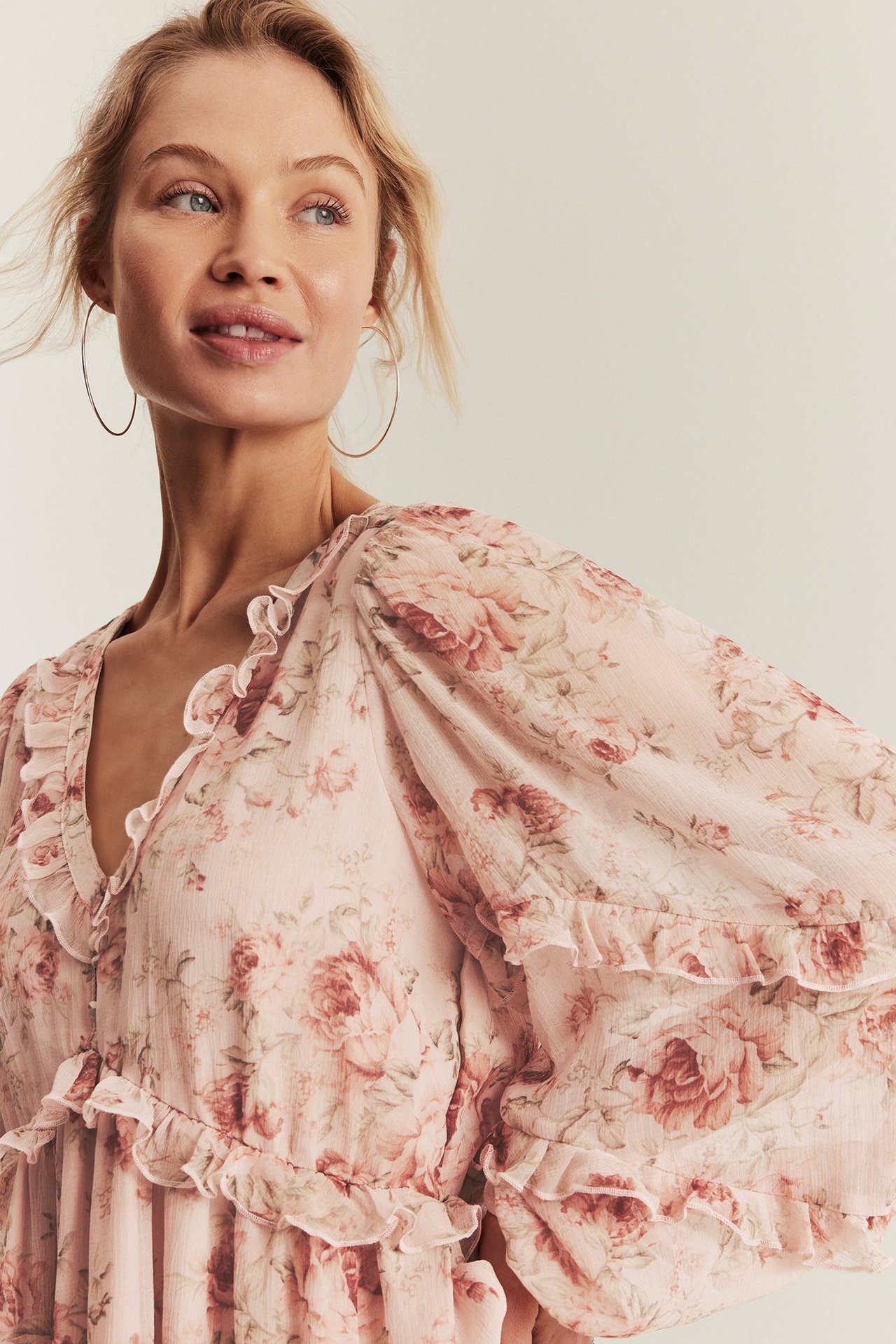 Close-up front view of a woman wearing a pink floral chiffon dress with ruffles and large silver hoop earrings.