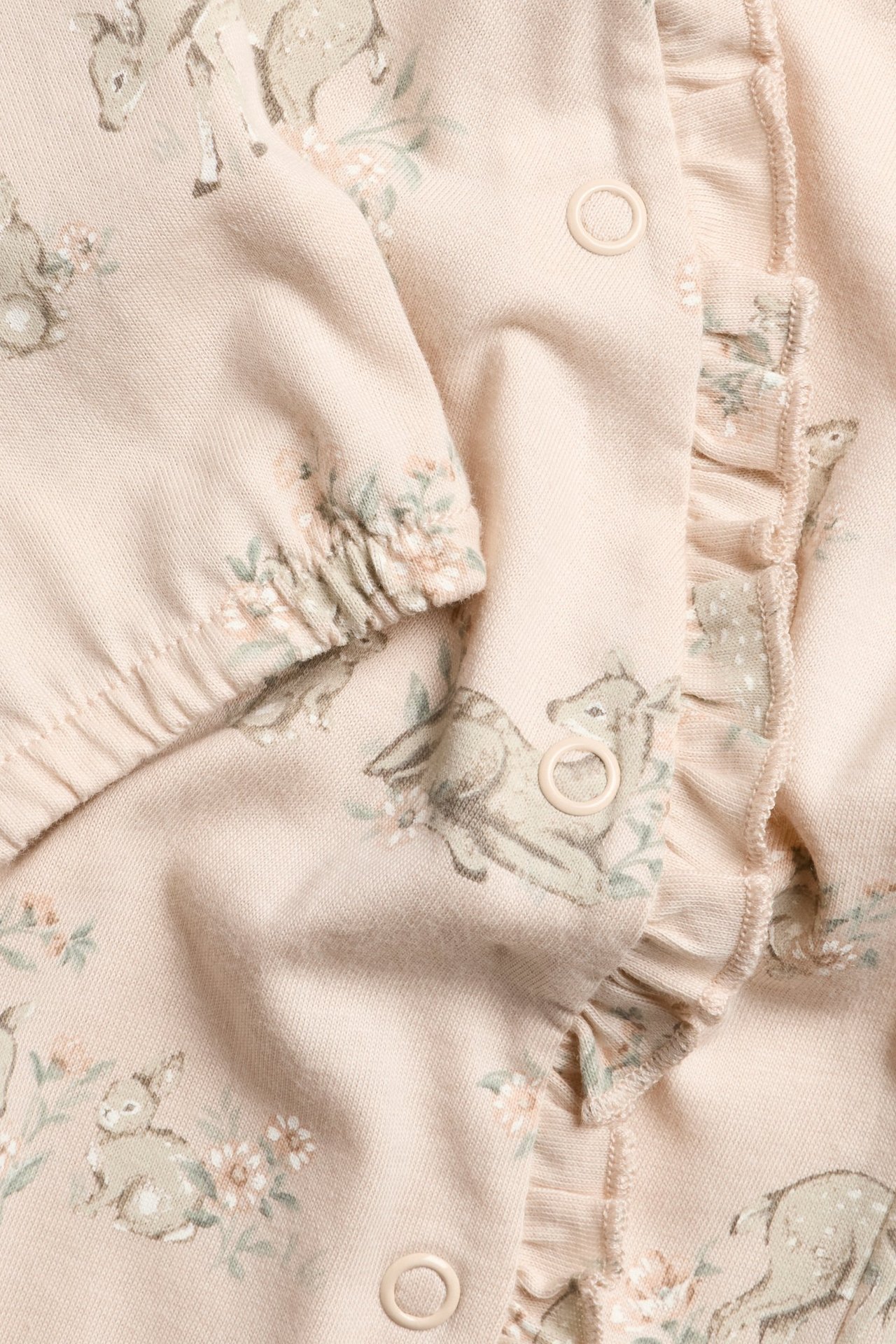 Nahaufnahme eines rosa Baby-Baumwolljersey-Stramplers mit Reh- und Blumenmuster, Rüschen und Druckknöpfen.