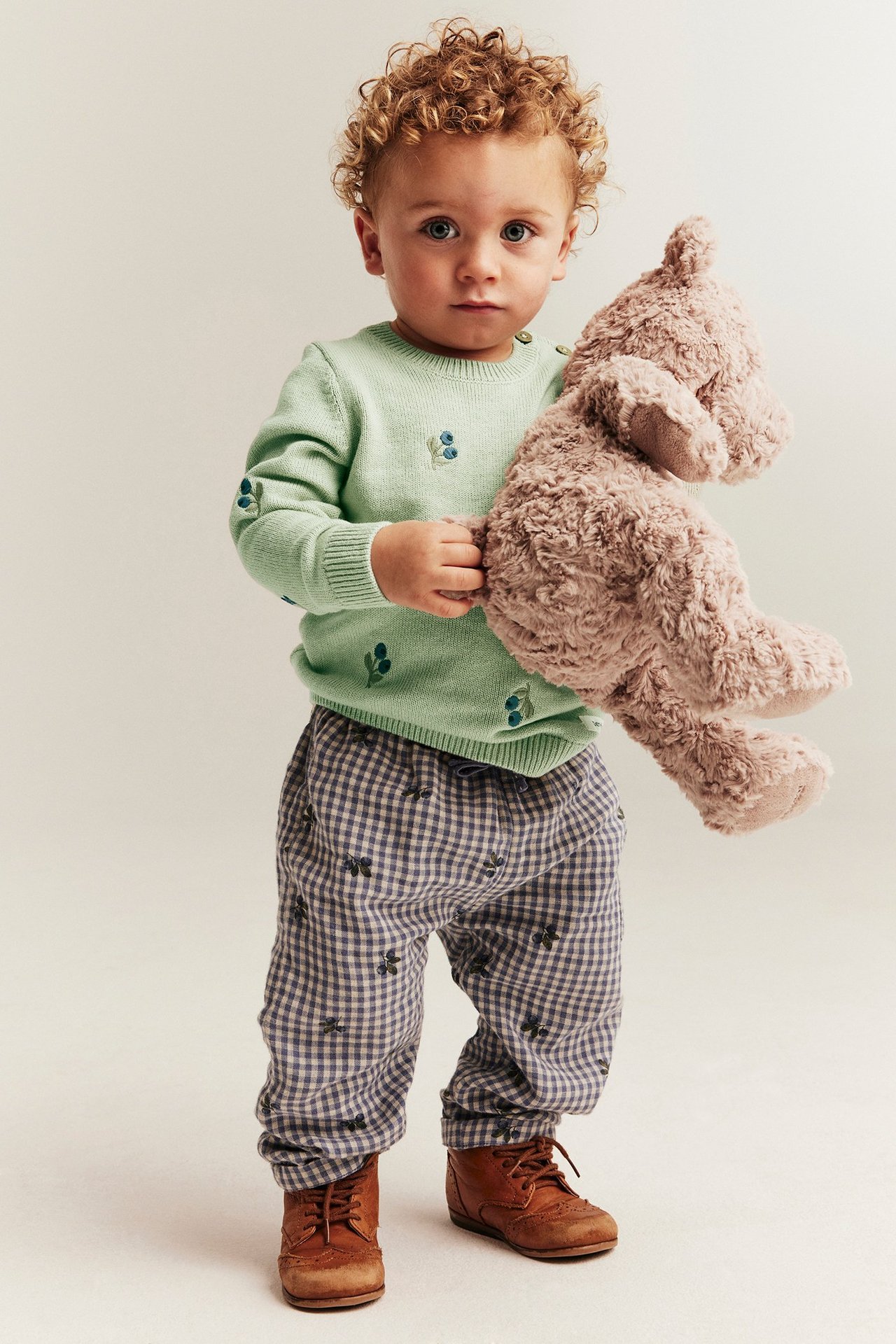 Front view of a baby in a light green embroidered sweater, blue checked pants, brown shoes, holding a plush toy.