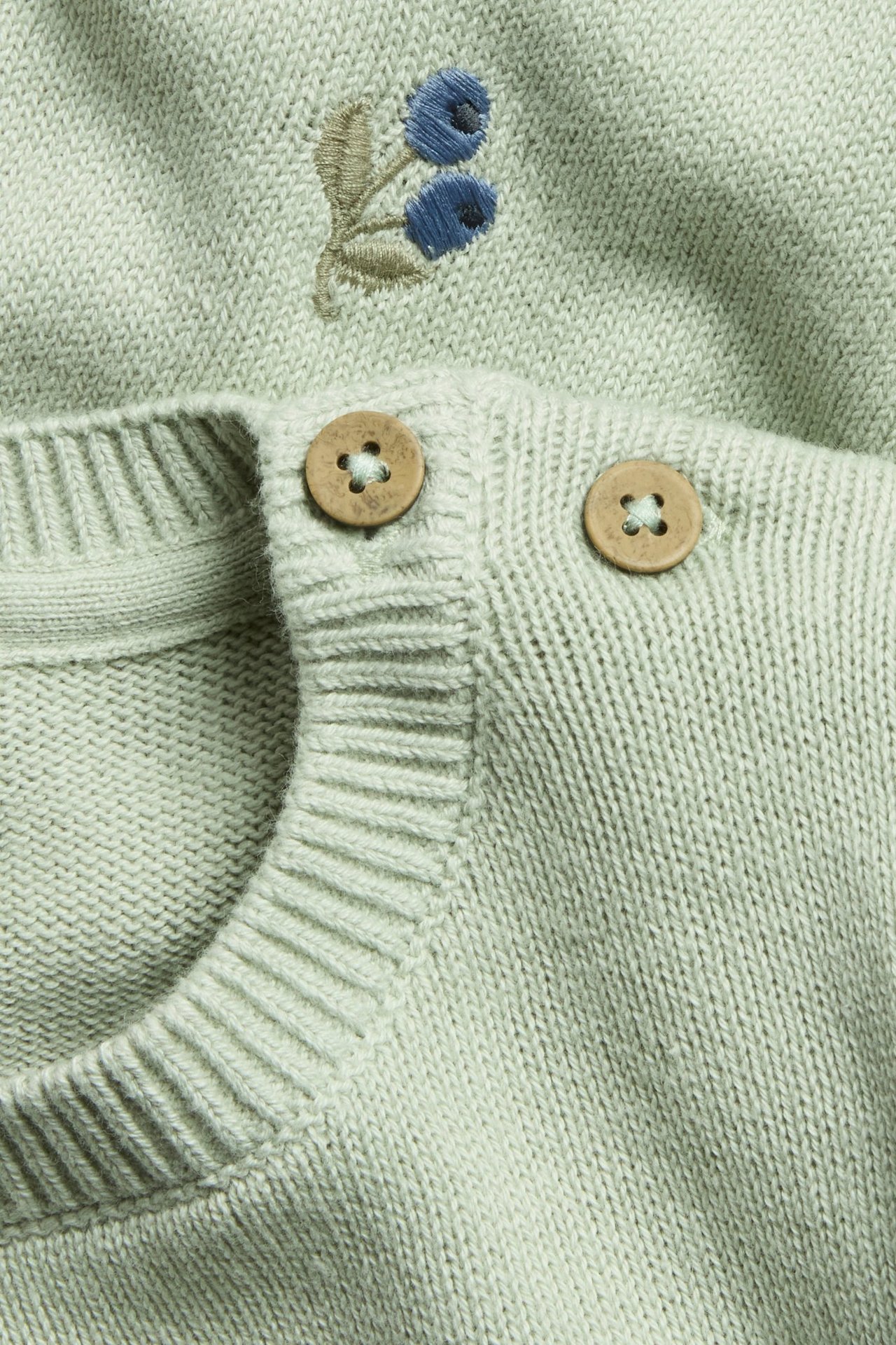 Nahaufnahme: Hellgrüner Jacquard-Strickpullover für Babys, mit blauer Beerenstickerei und zwei Holzschulterknöpfen.