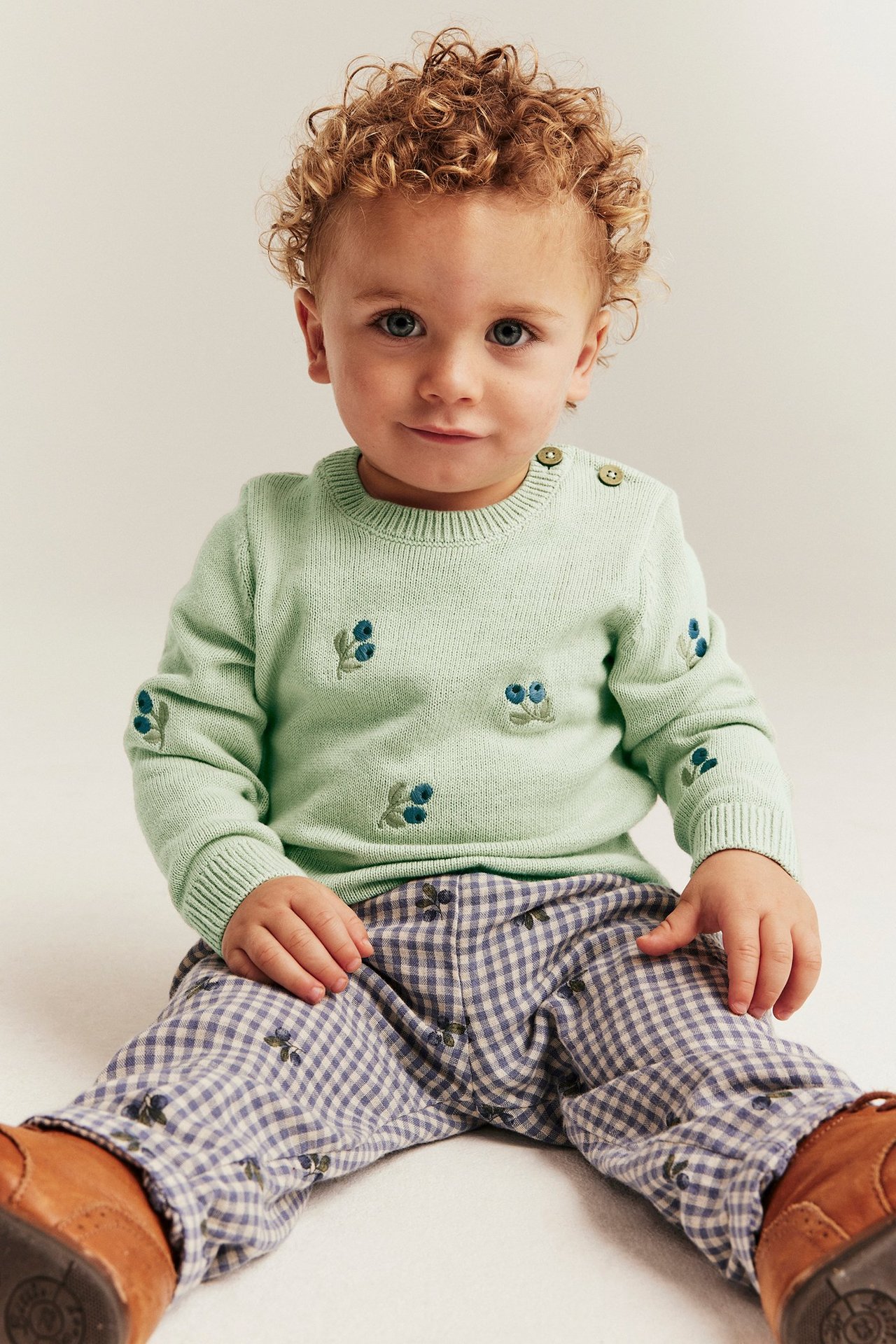 Front view: Baby in light green cotton sweater with blueberry embroidery, shoulder buttons, gingham pants, brown shoes.