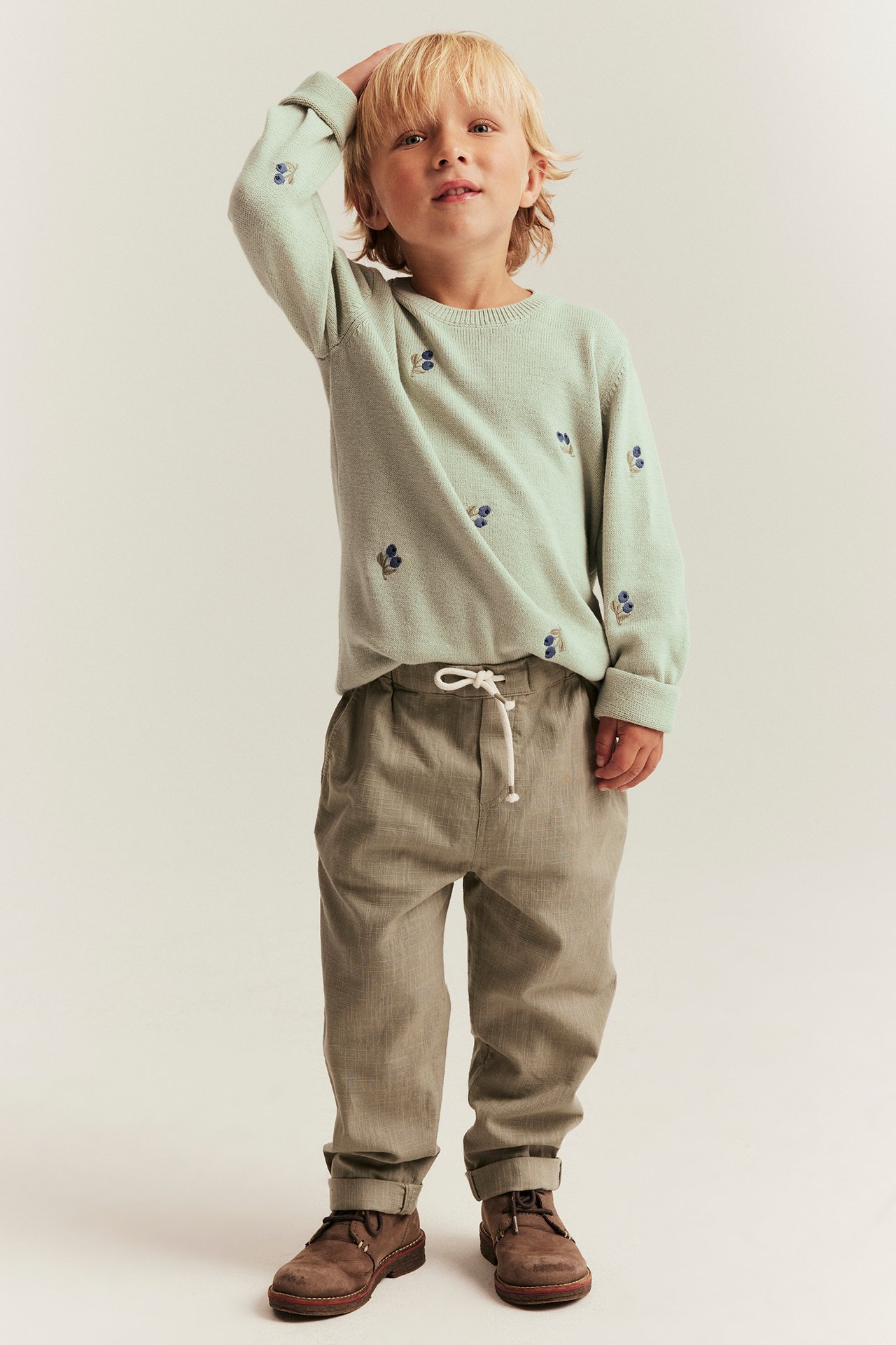 Front view kid in a light green jacquard sweater with blue embroidery, beige trousers, and brown shoes.