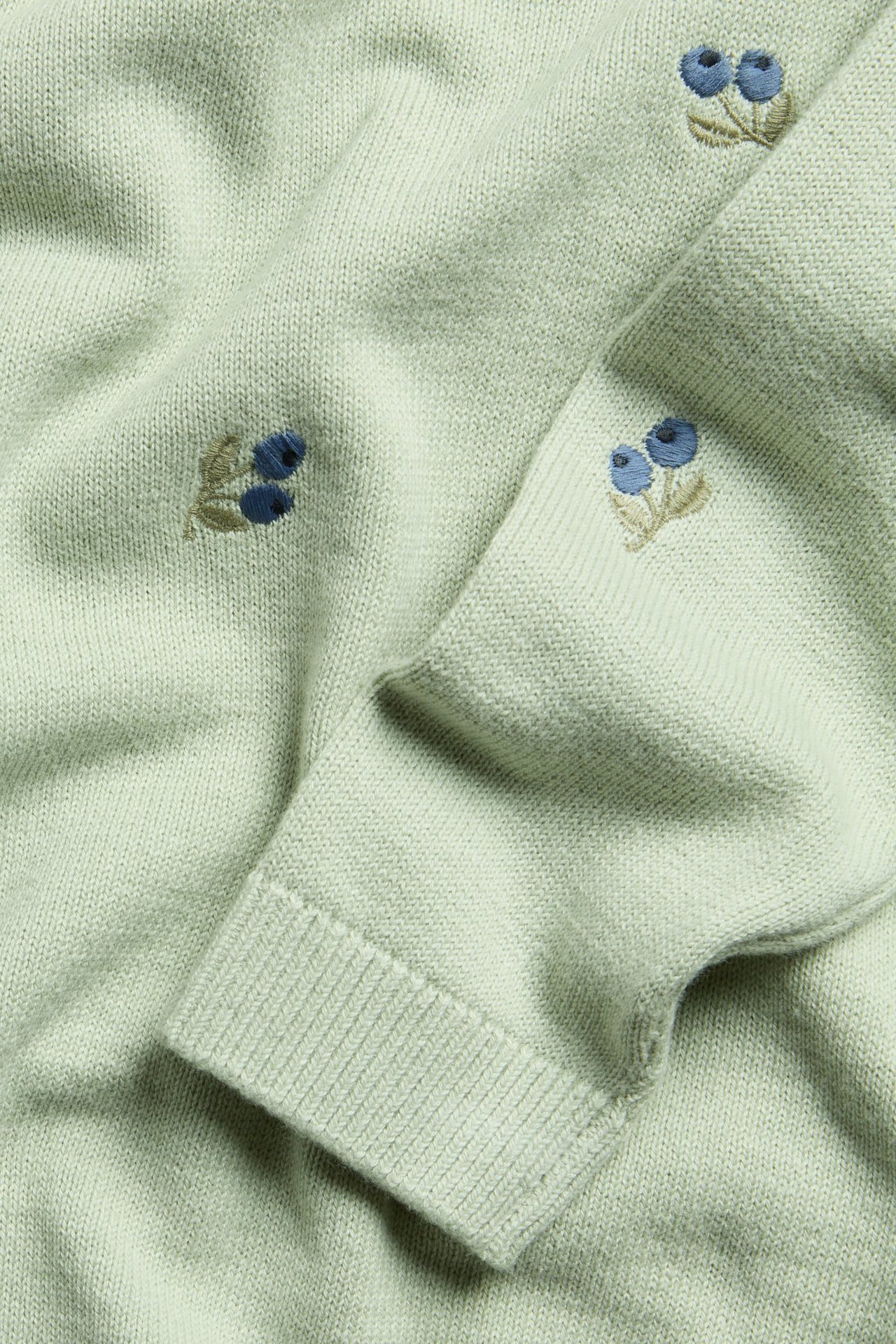 Nahaufnahme eines hellgrünen jacquardgestrickten Baumwollpullovers mit blauen, bestickten Heidelbeermustern und Rippenbündchen.