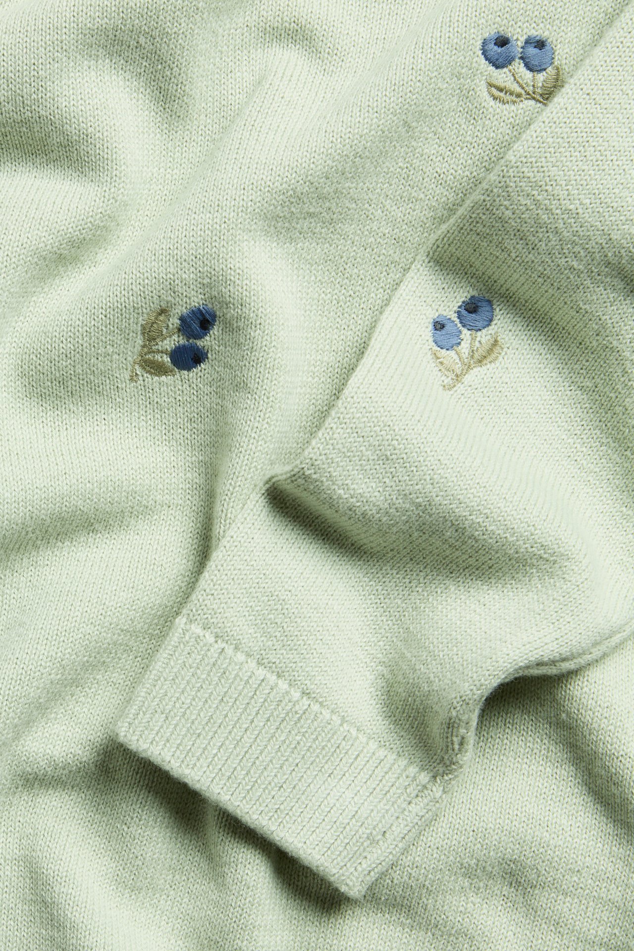 Close-up of a pale green jacquard-knitted cotton sweater with blue blueberry embroidery and ribbed cuffs.
