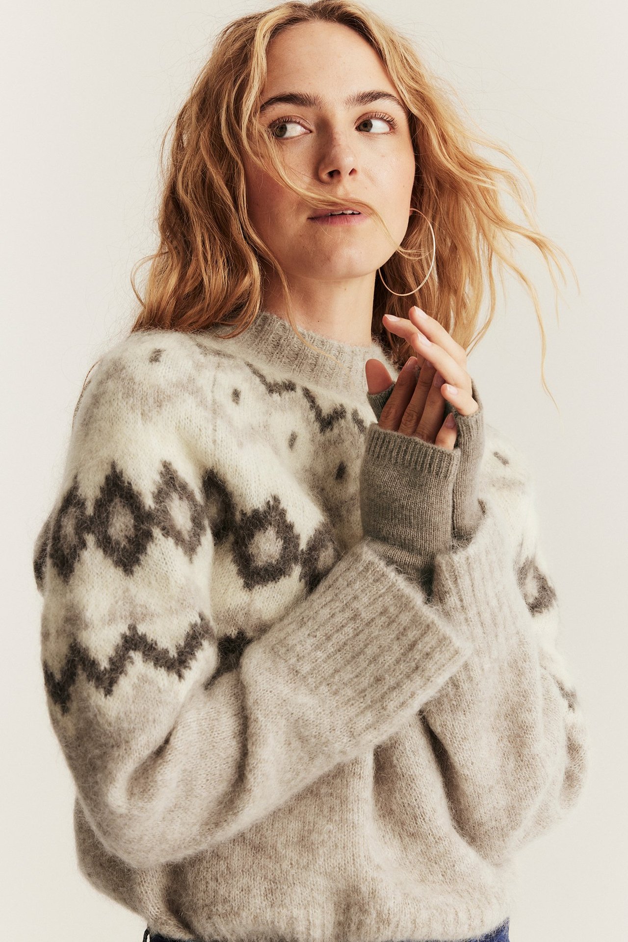 Nahaufnahme einer Frau mit beigemelierten Woll-Kaschmir-Handgelenkwärmern über einem gemusterten grauen Pullover und goldenen Creolen.